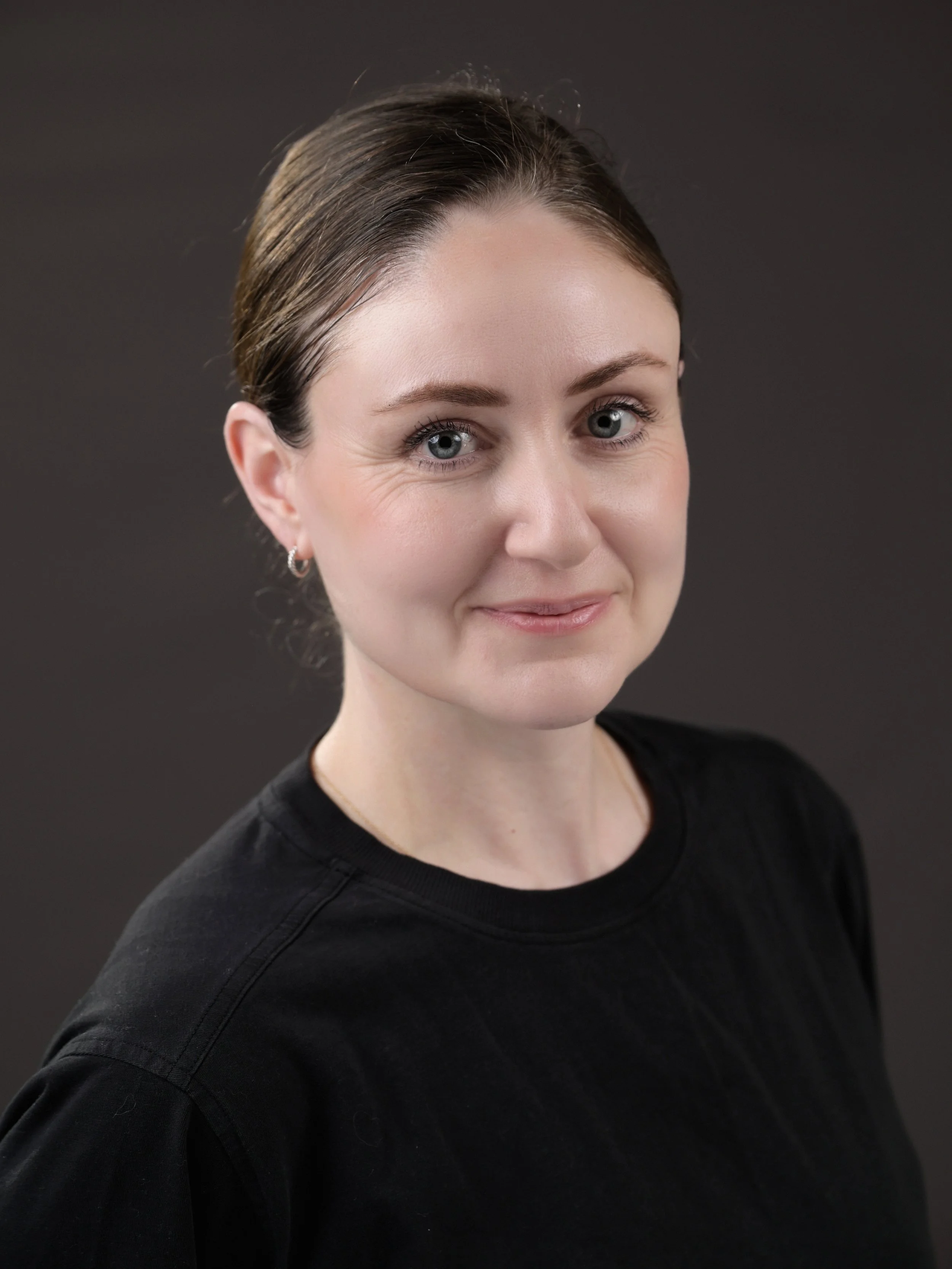 A young woman with fair skin, blue eyes, dark brown hair styled in a bun, wearing earrings and a black shirt, smiling softly against a dark background.