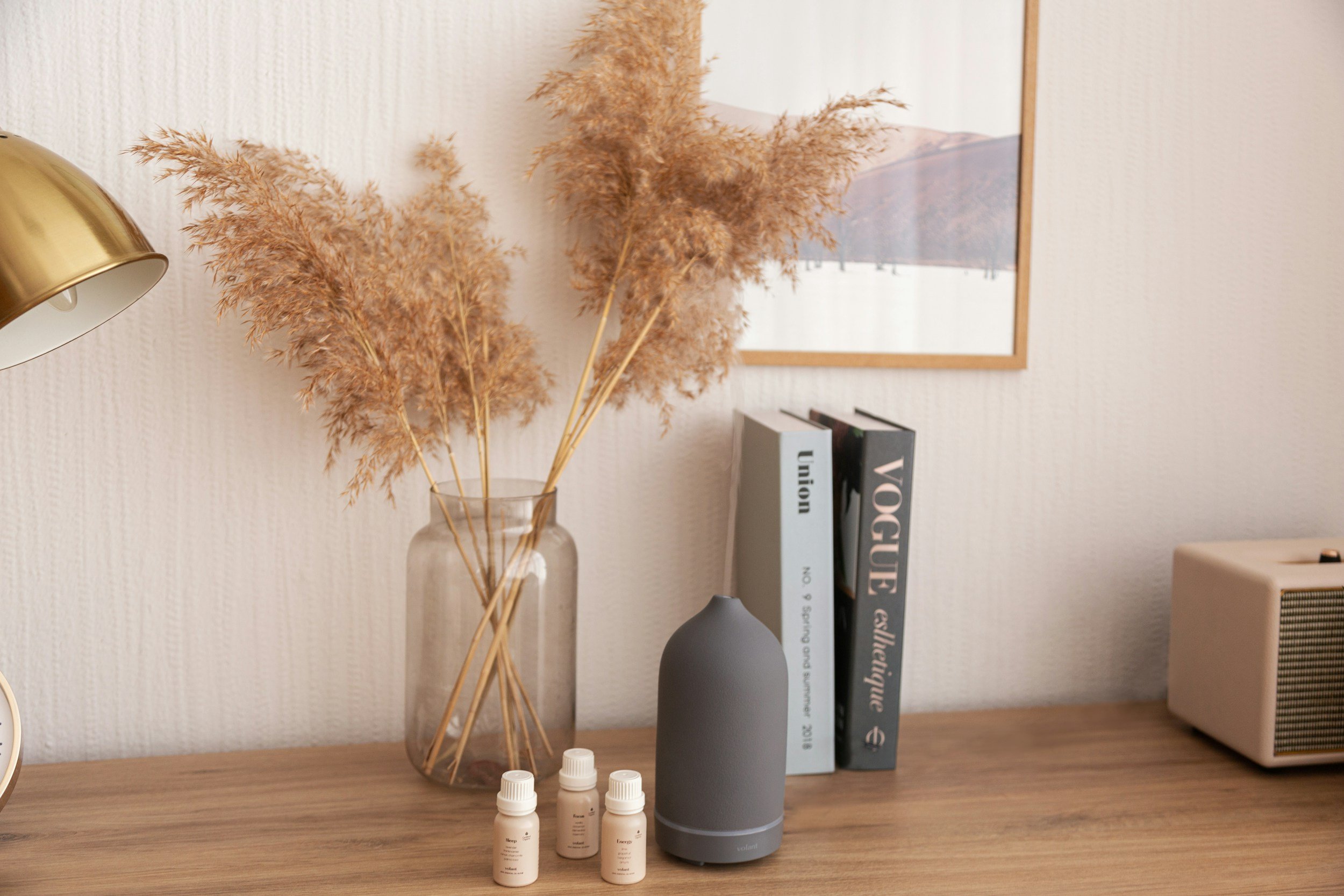 A wooden table with a gray essential oil diffuser, four small bottles of essential oils, and a clear glass vase holding dried pampas grass. In the background, a framed landscape photograph and a stack of books are visible, along with a brass-colored desk lamp.
