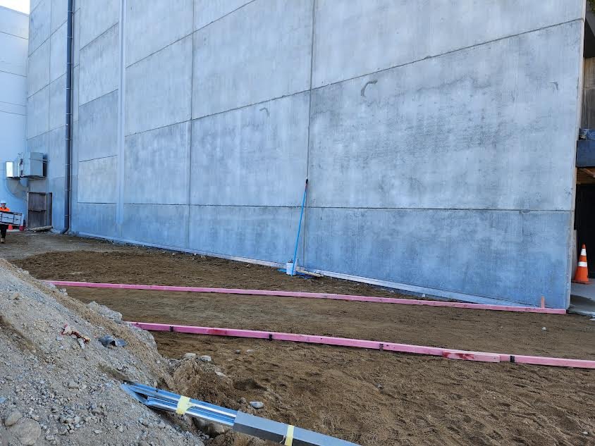 Construction site with a large concrete wall, dirt ground, pink excavation barriers, and construction cones.