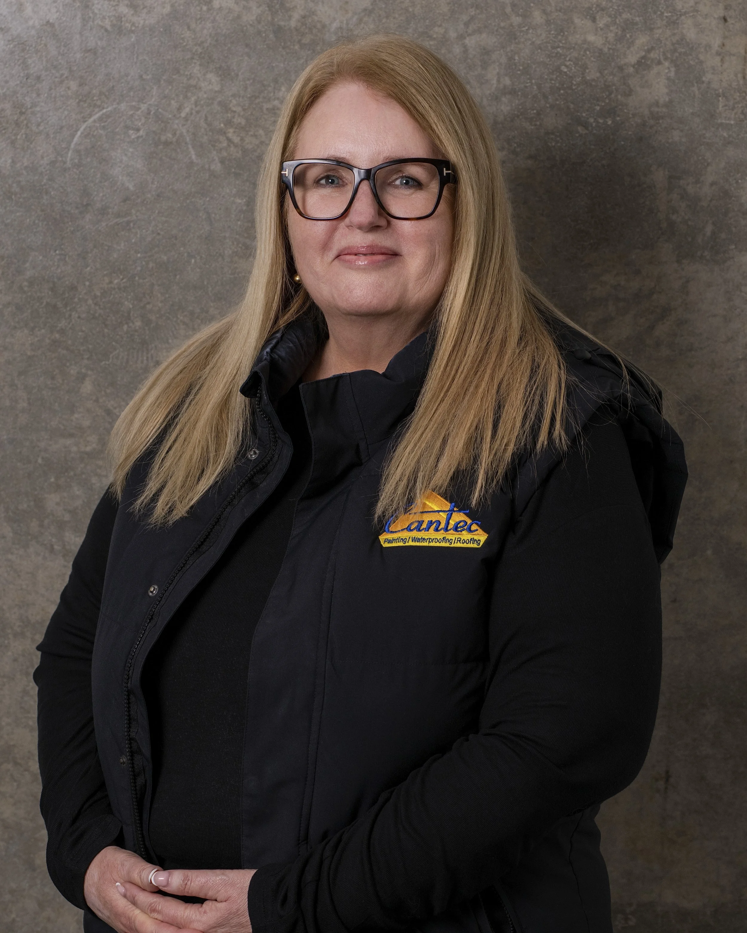 A woman with long blonde hair, wearing large eyeglasses and a black jacket with a patch that says 'Lancel Painting Waterproofing Roofing', is standing against a gray textured background. She is looking at the camera with a slight smile and has her hands folded in front of her.