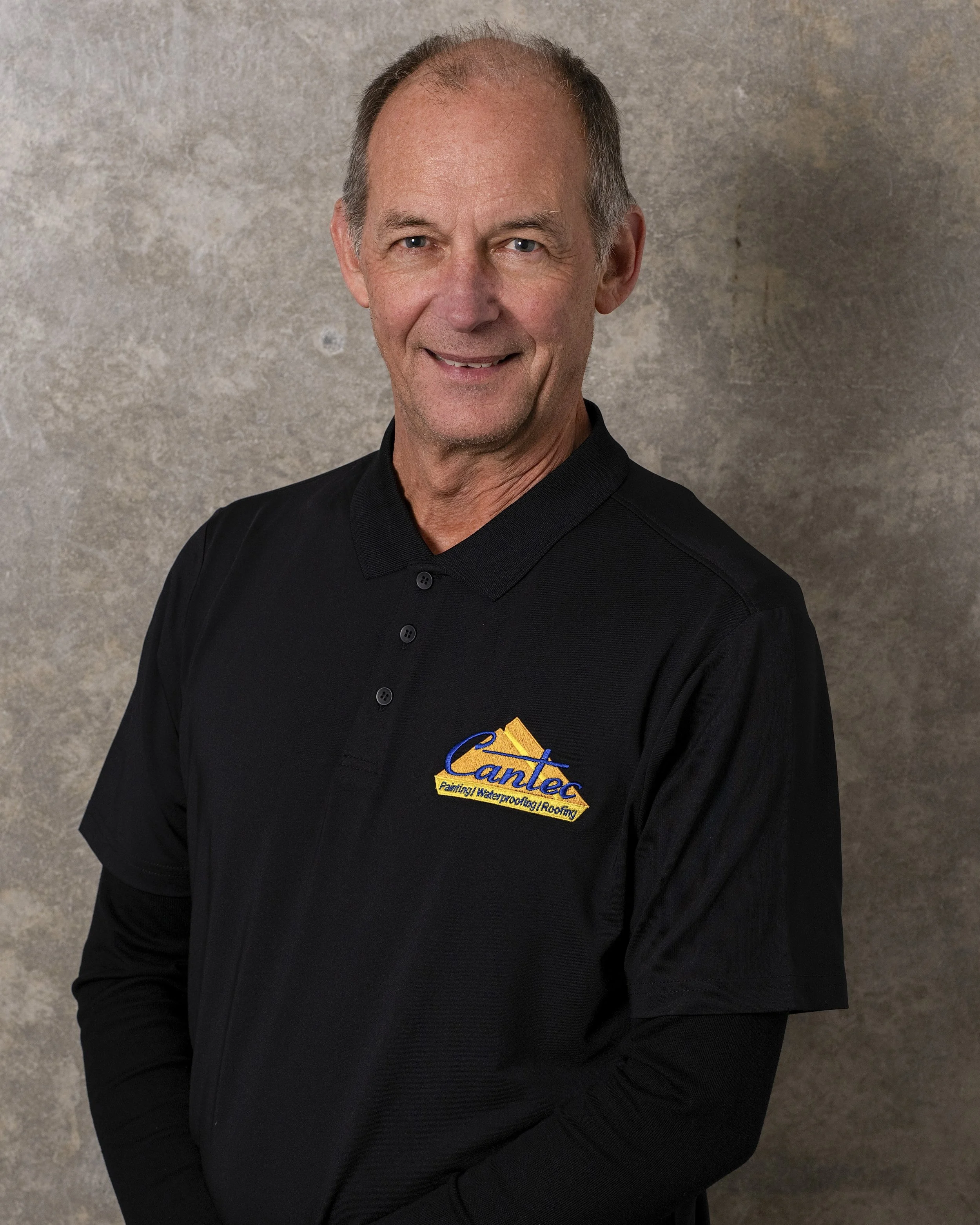 A middle-aged man with short hair, dressed in a black shirt with the logo of Cantec Painting, Waterproofing, Roofing embroidered on it, standing against a textured gray background.