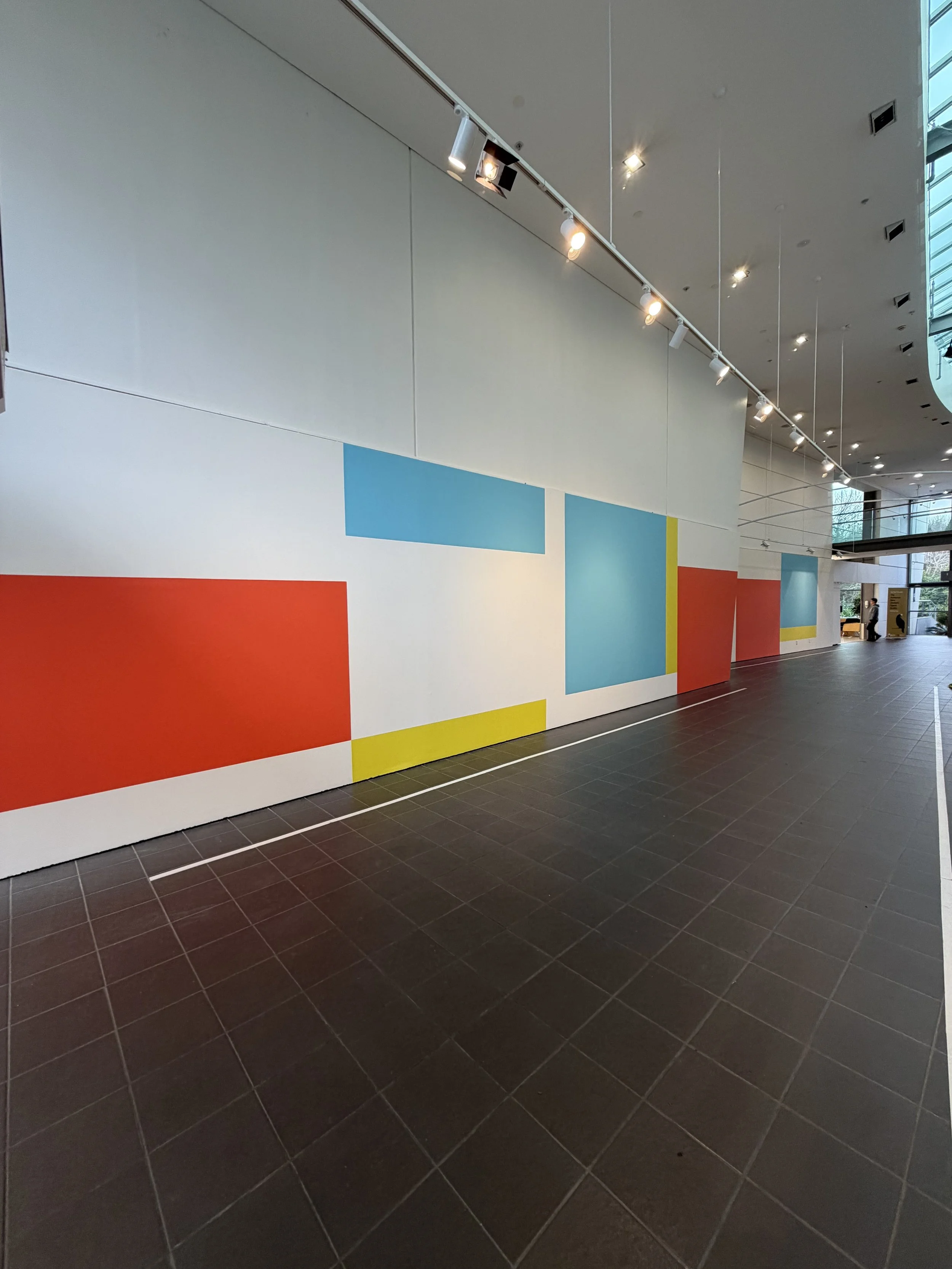 Interior view of a modern building with a large abstract mural on the wall featuring colorful blocks of red, blue, yellow, light blue, and white. The ceiling has track lighting with spotlights, and the floor is made of dark gray tiles. In the background, windows and a person are visible.