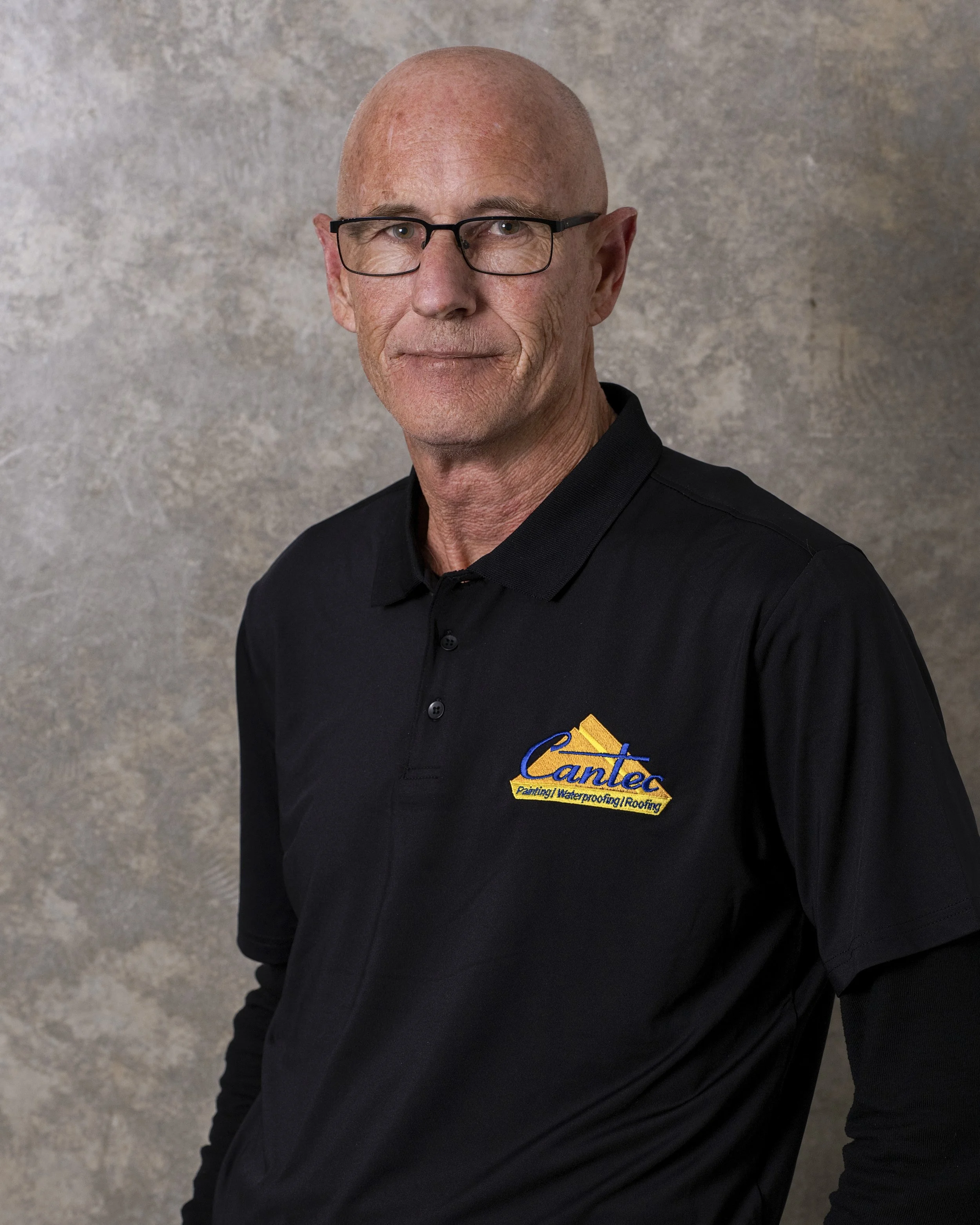 A bald man with glasses wearing a black collared shirt with a company logo that reads, "Cantex Painting Waterproofing Roof". He is standing in front of a neutral textured grey background.