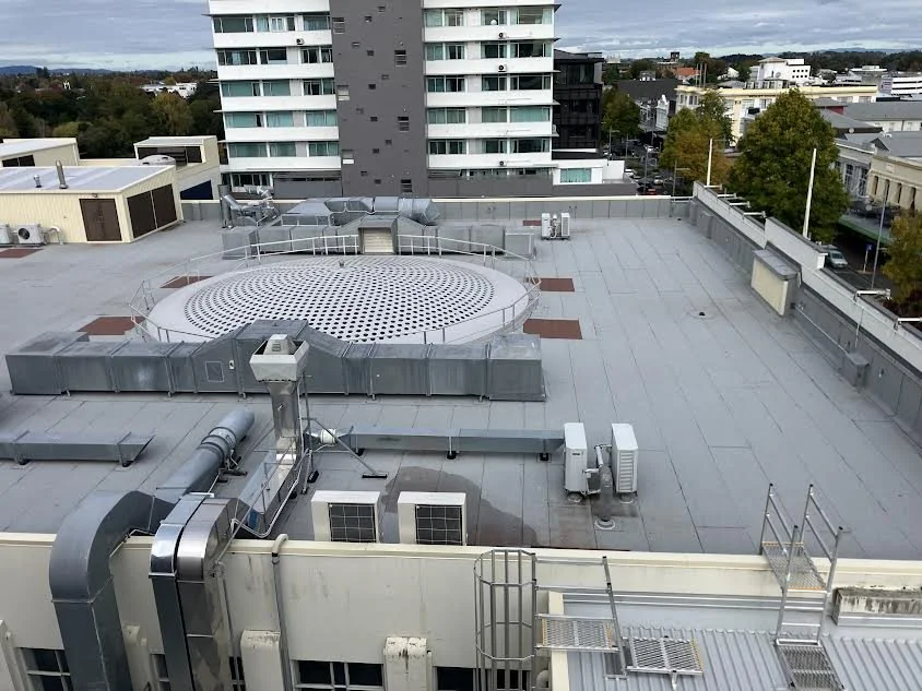 View of a flat rooftop with equipment, vents, and ductwork, with a high-rise building and trees in the background.