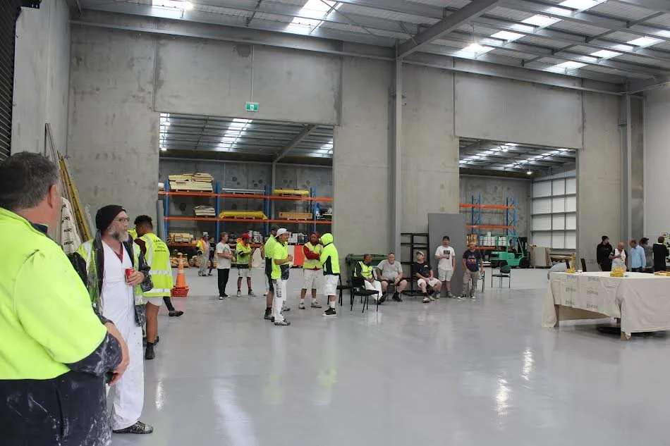 People inside a spacious warehouse or industrial building, some wearing safety vests, others sitting on chairs, with tables, equipment, and shelving units around.