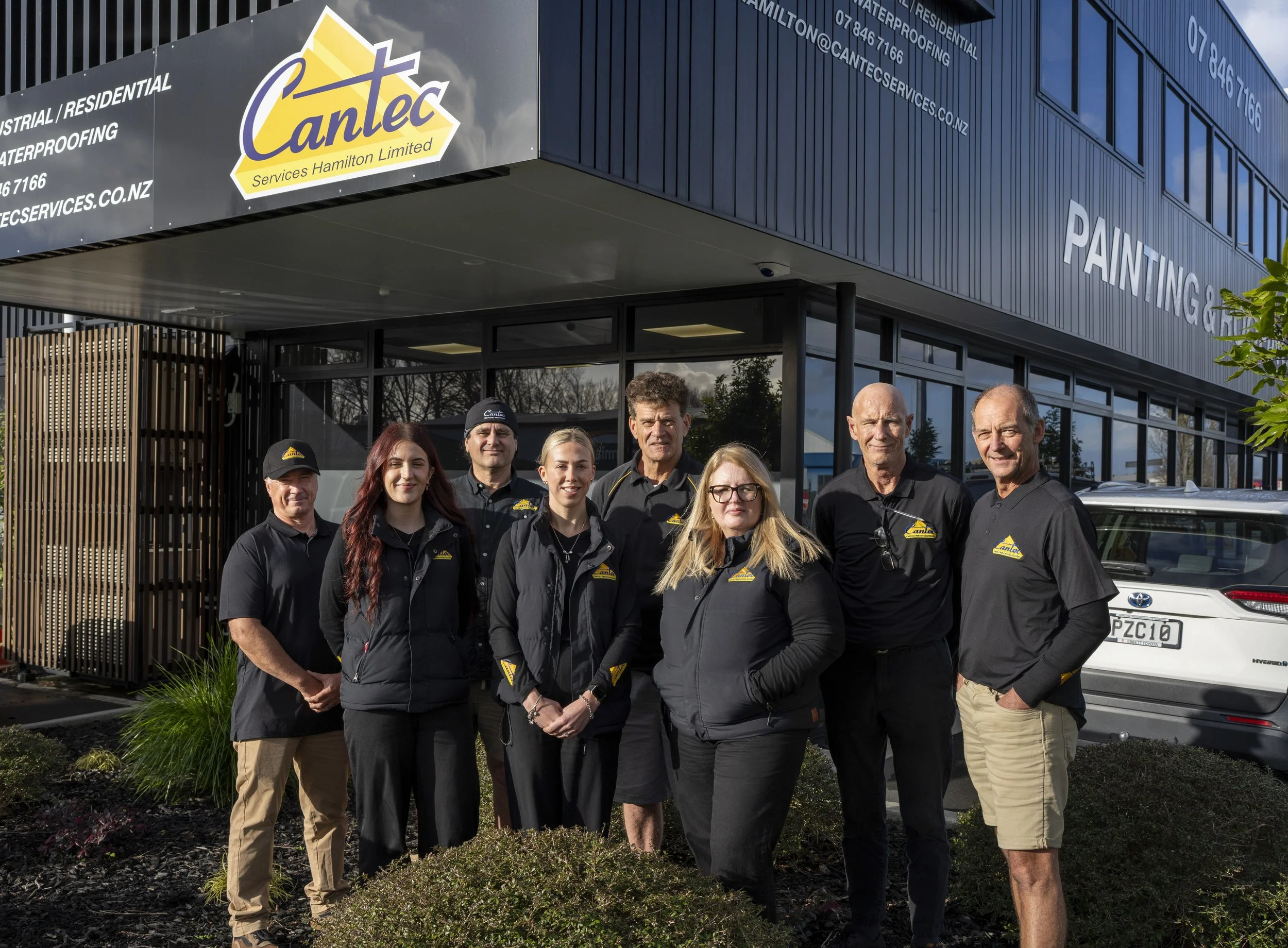 A group of nine people standing outside a commercial building with signage for Canlec Services Hamilton Limited, a waterproofing business. The group appears to be employees or staff members, dressed in black shirts with the company logo. The building has large windows and a sign that reads 'PAINTING.'