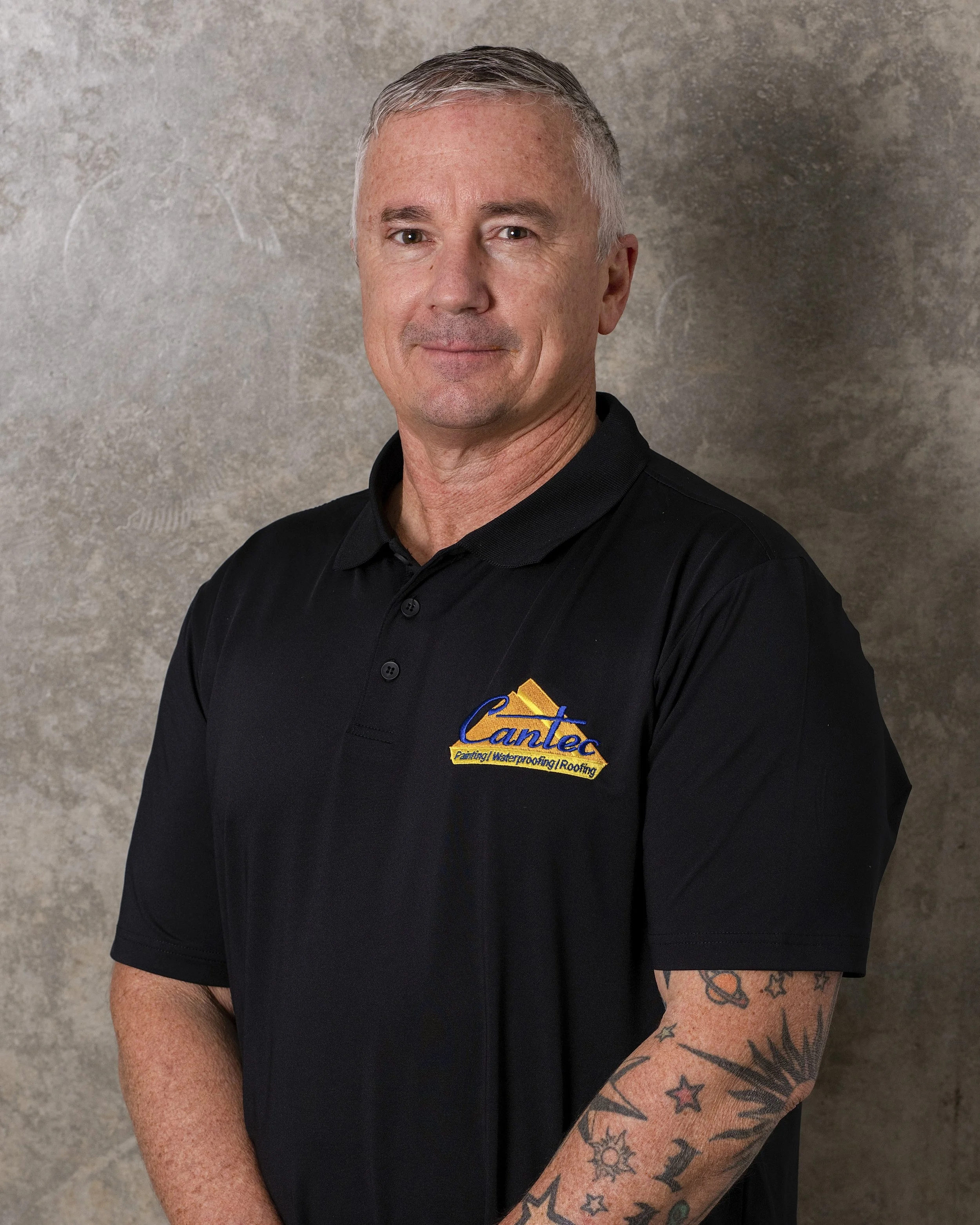 Portrait of a middle-aged man with short gray hair, wearing a black collared shirt with a logo on the chest, standing against a textured gray wall.