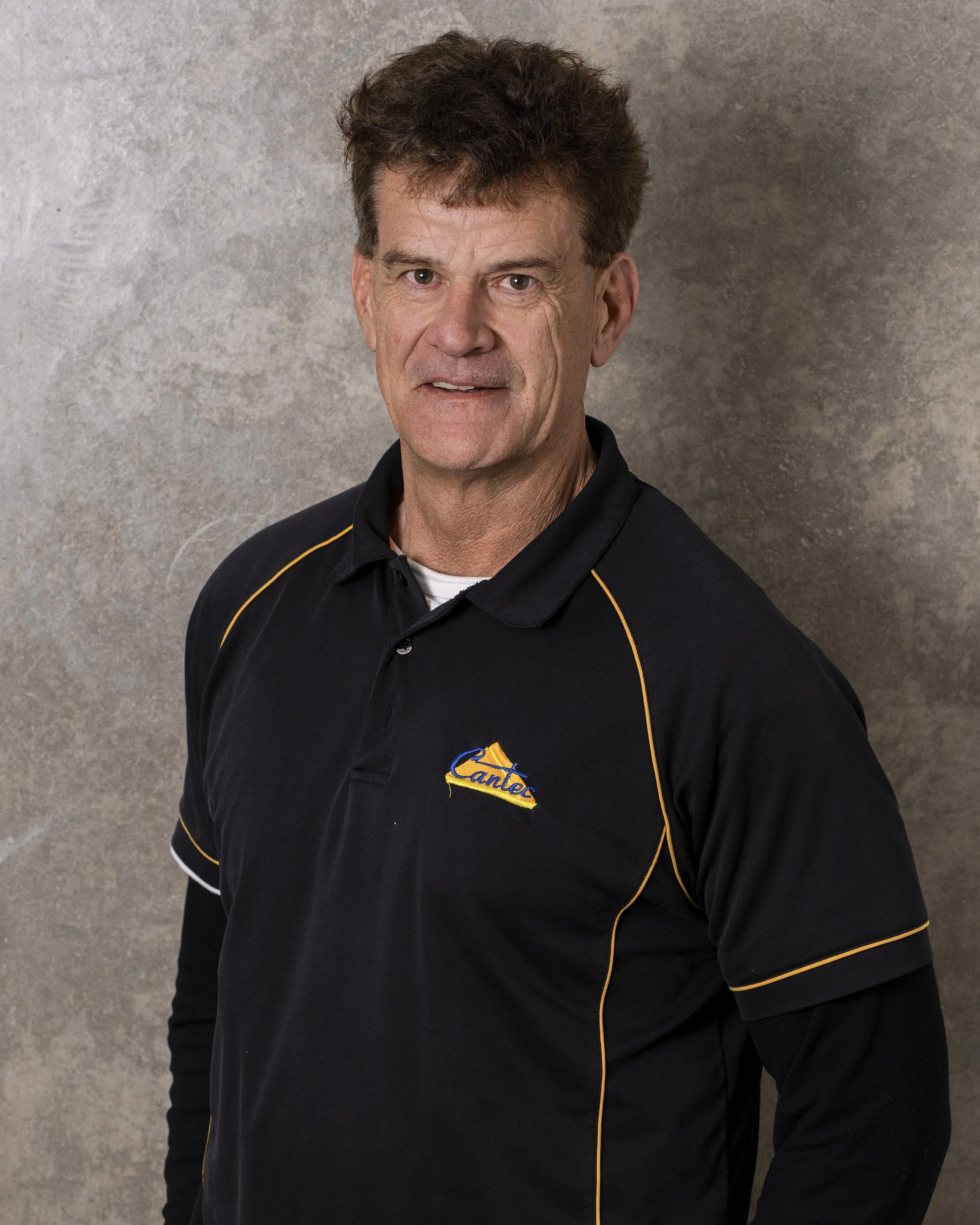 A man with dark, curly hair wearing a black sports shirt with yellow piping and an embroidered logo that reads 'Cante' or 'Cantex' on the chest, standing in front of a gray textured background.