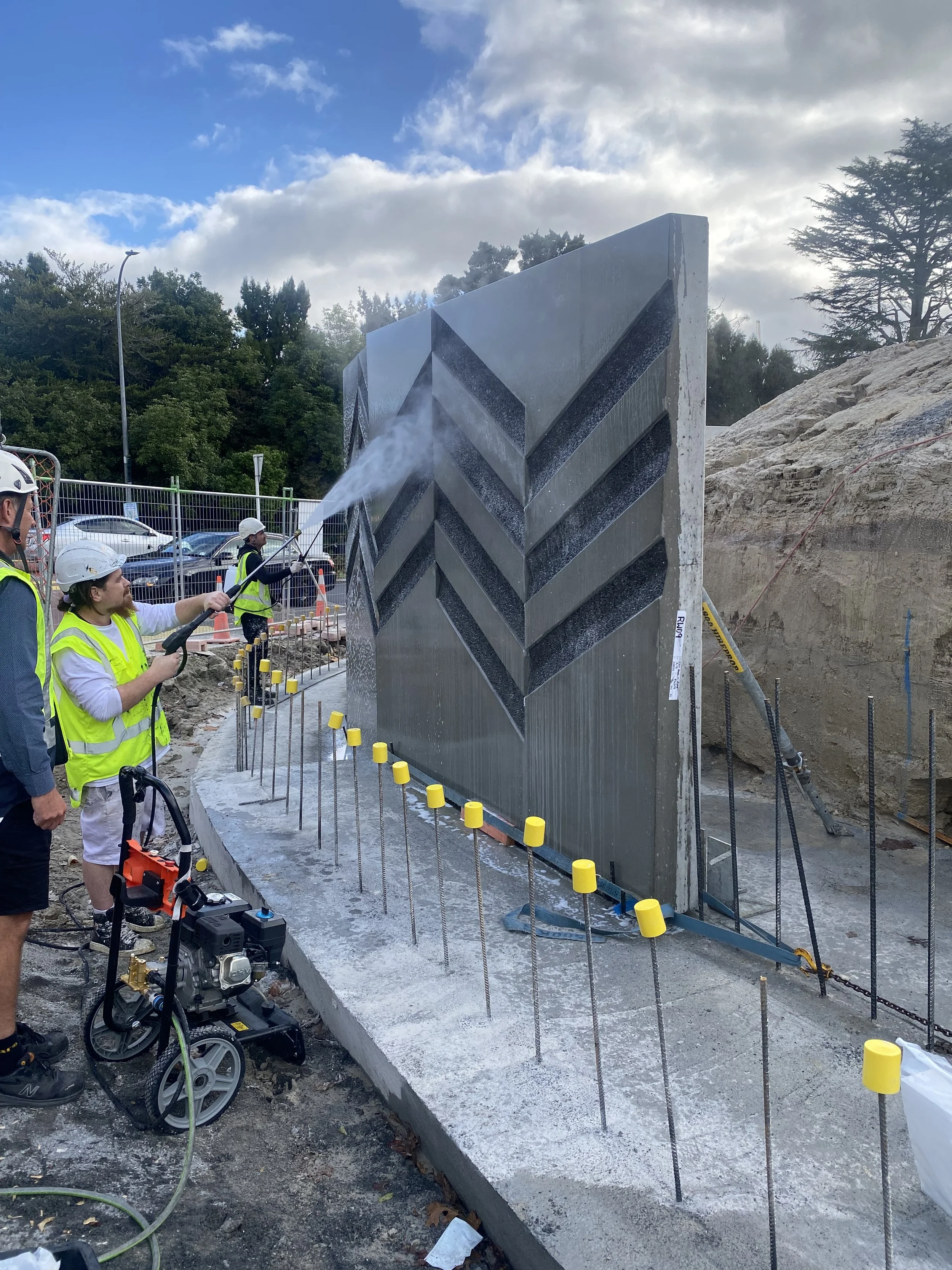 Workers in safety helmets and vests pressure washing a large decorative concrete wall during construction, with fencing and parked cars in the background.