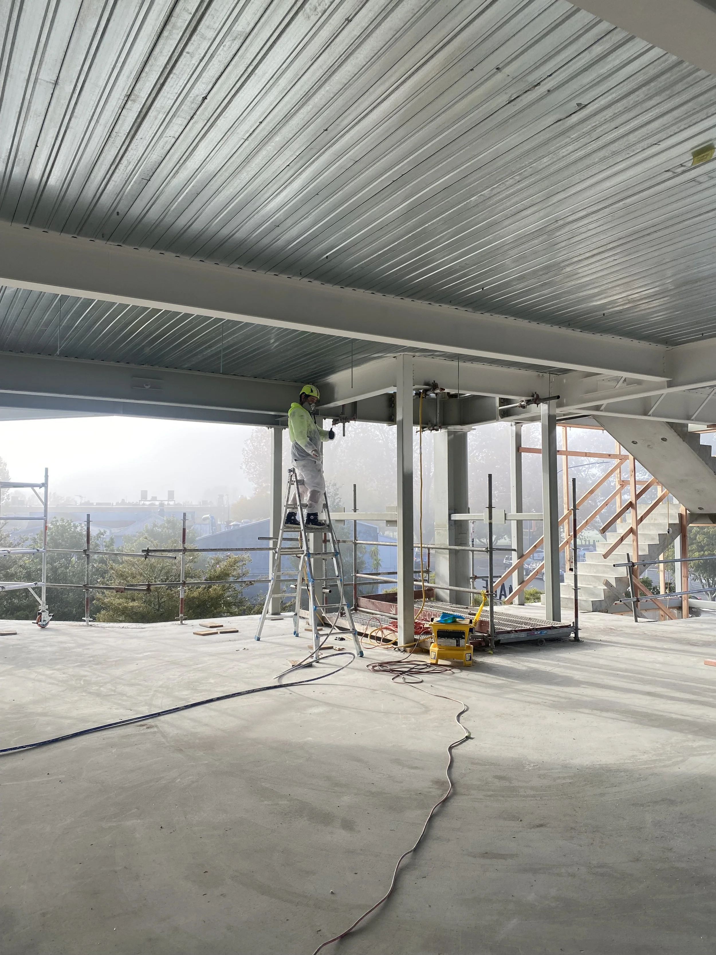Construction worker on ladder working on ceiling inside a building with scaffolding, metal beams, and stairs visible.