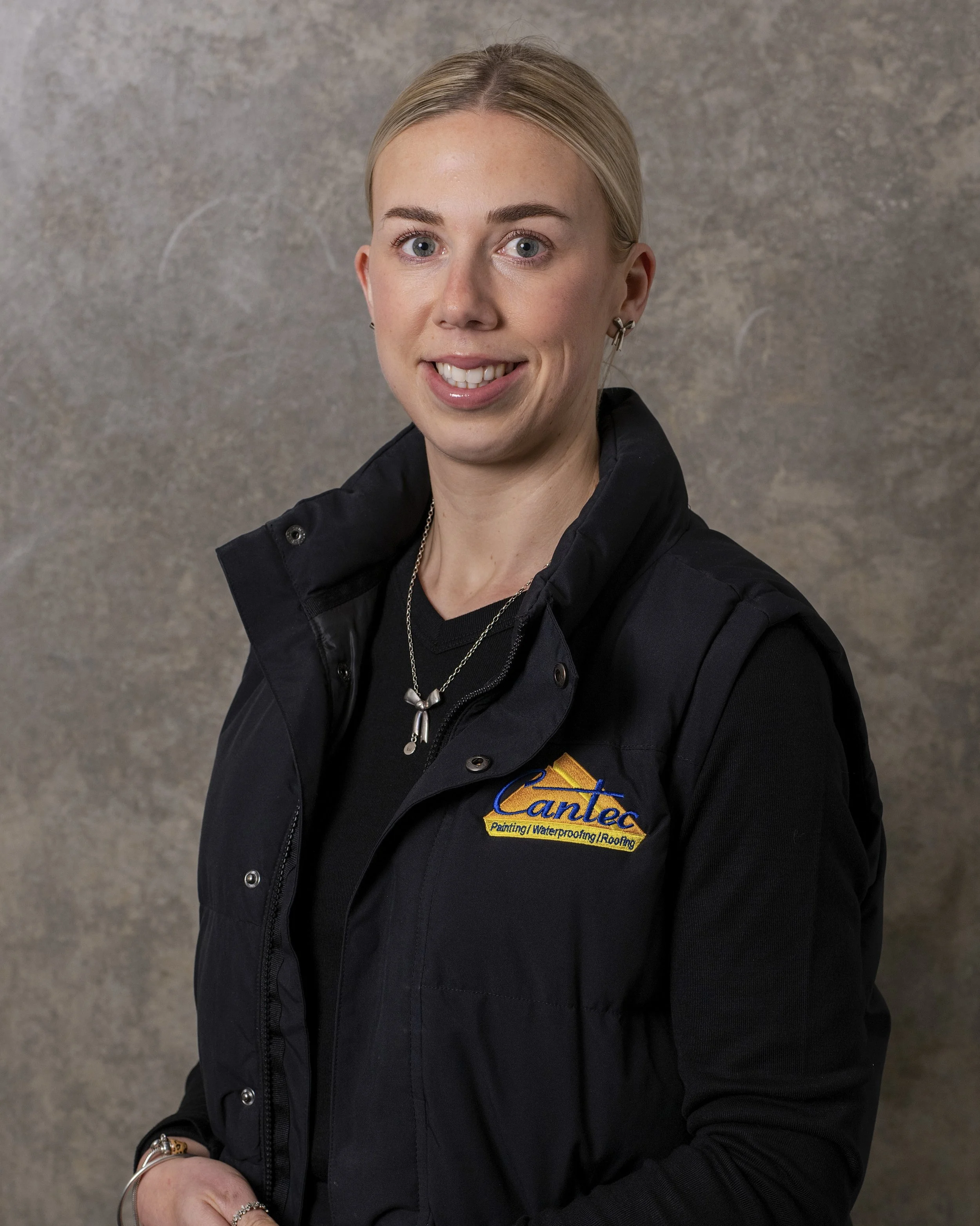A woman with blonde hair, blue eyes, and earrings smiling at the camera, wearing a black jacket with an embroidered logo that reads 'Cantos Painting Waterproofing Roofing' against a gray background.