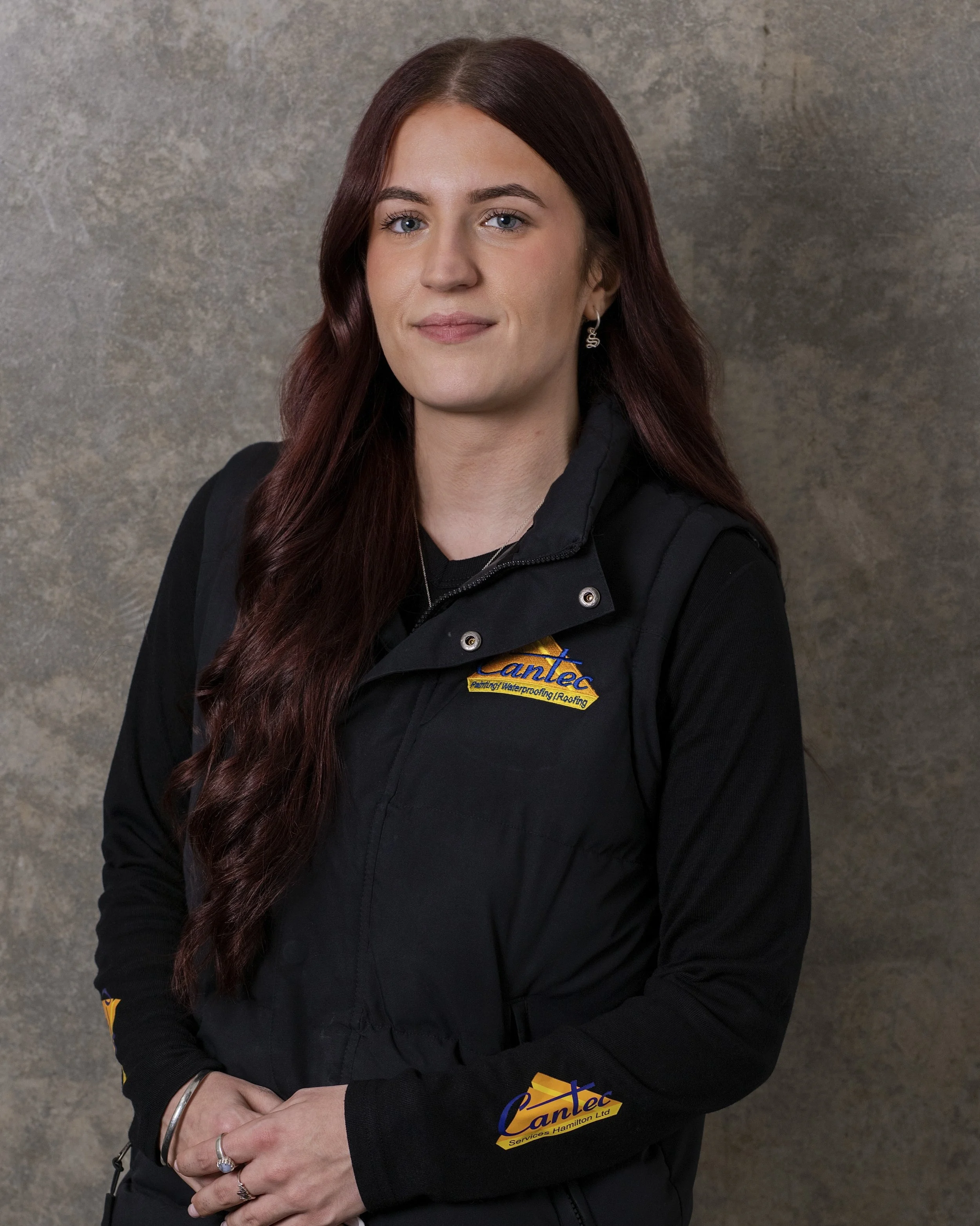 A woman with long red hair and blue eyes wearing a black jacket with a Cantex logo standing against a textured gray background.