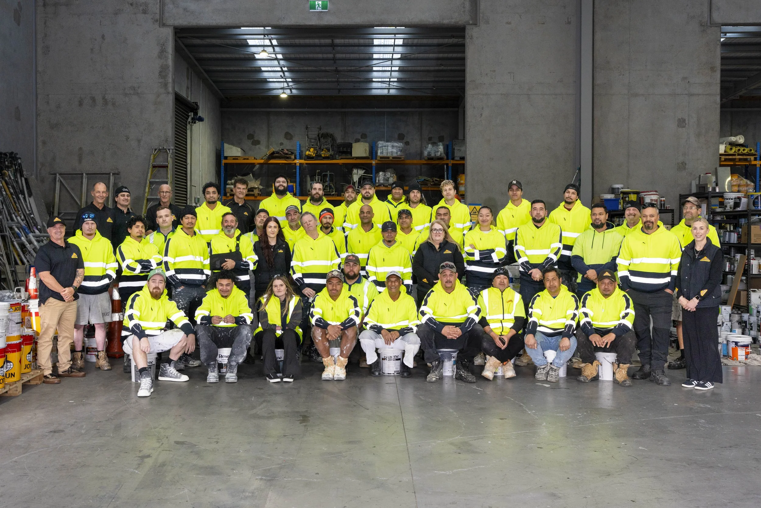 Group of workers in high-visibility jackets inside a warehouse, standing and sitting in front of shelves with supplies and tools.