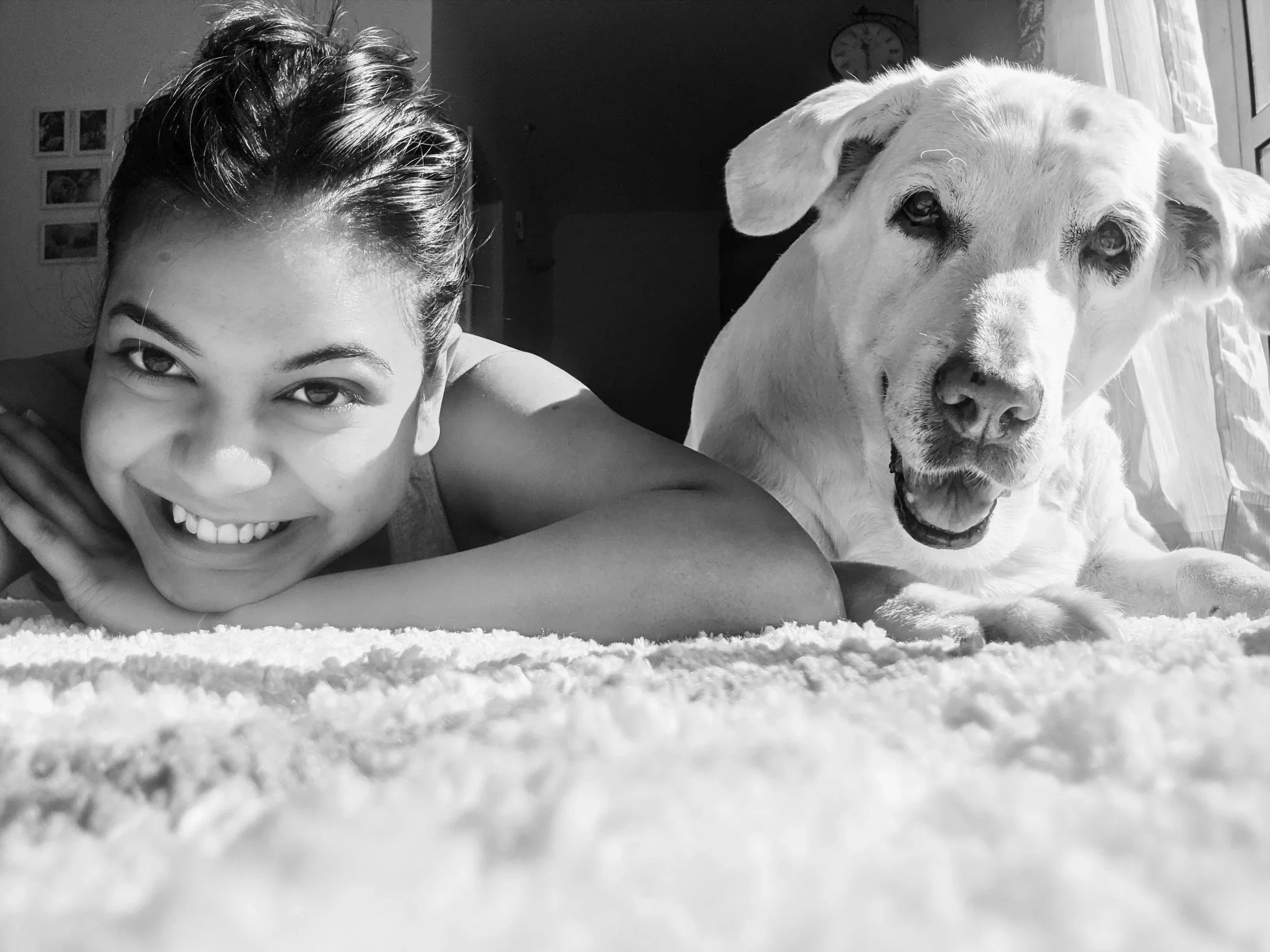 A woman lying on the floor with her head resting on her arm, smiling, and a large dog sitting next to her. The photo is in black and white.