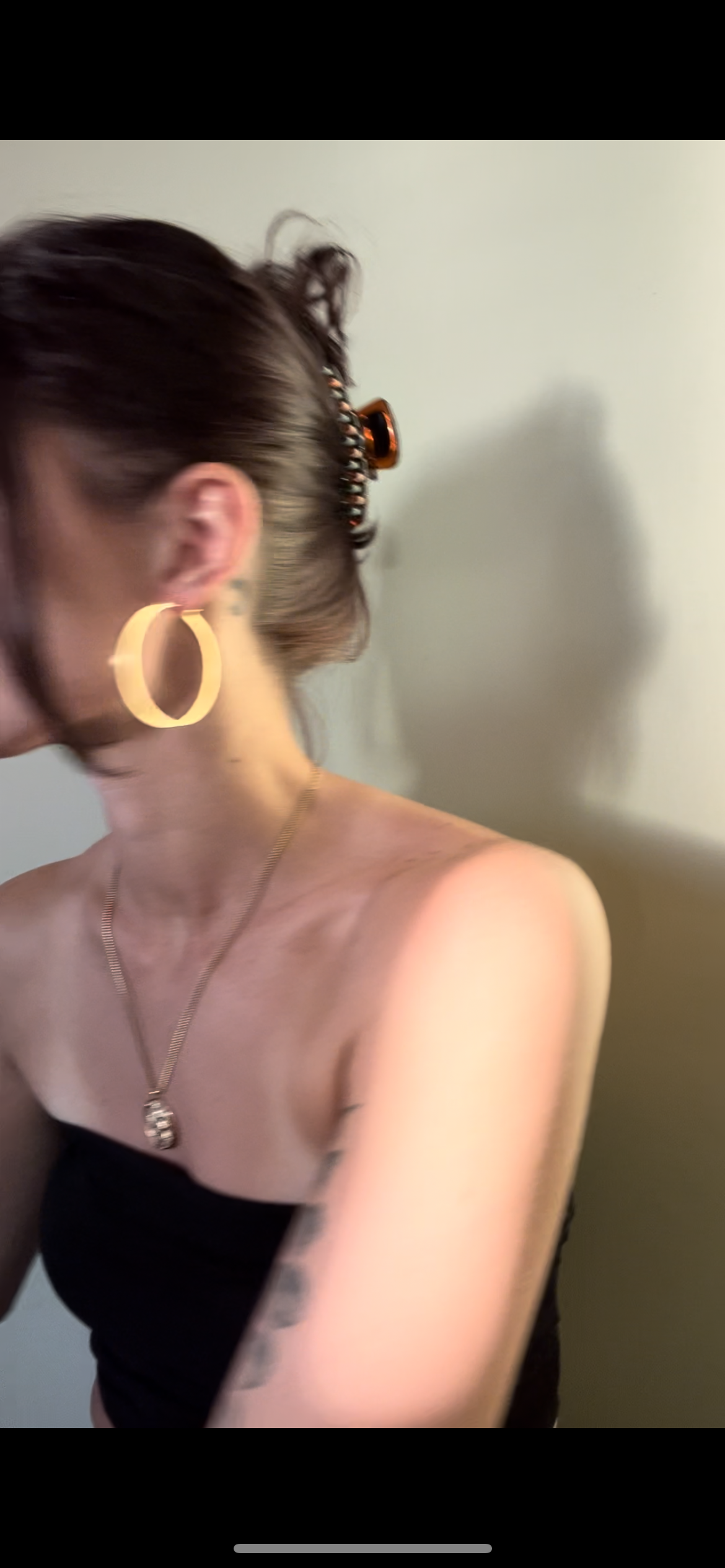 A woman with short brown hair, wearing large hoop earrings, a gold necklace with a pendant, and a black strapless top, standing against a light-colored wall, with her face turned away.