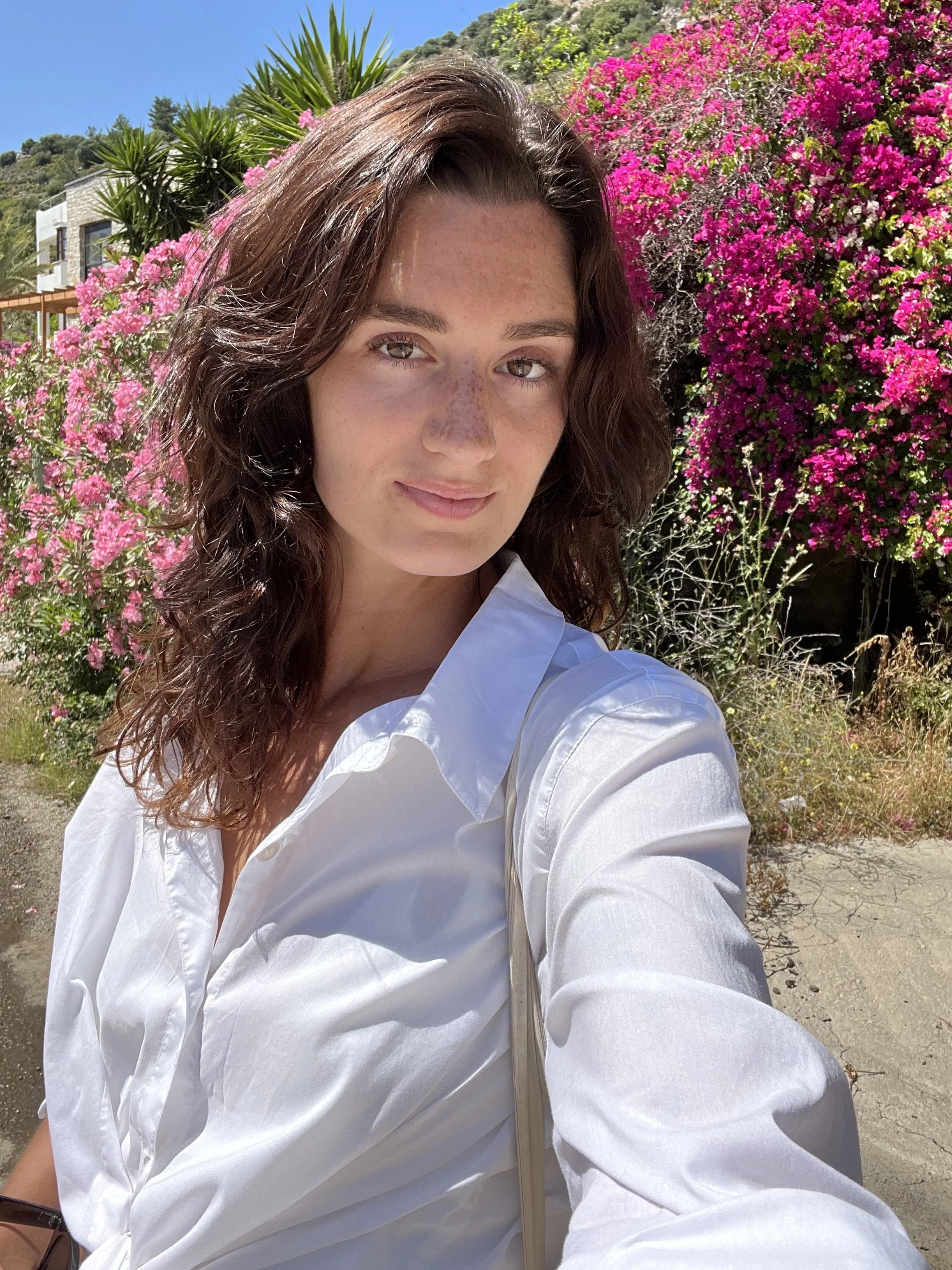 A young woman taking a selfie outdoors with pink and purple flowering bushes in the background on a sunny day.