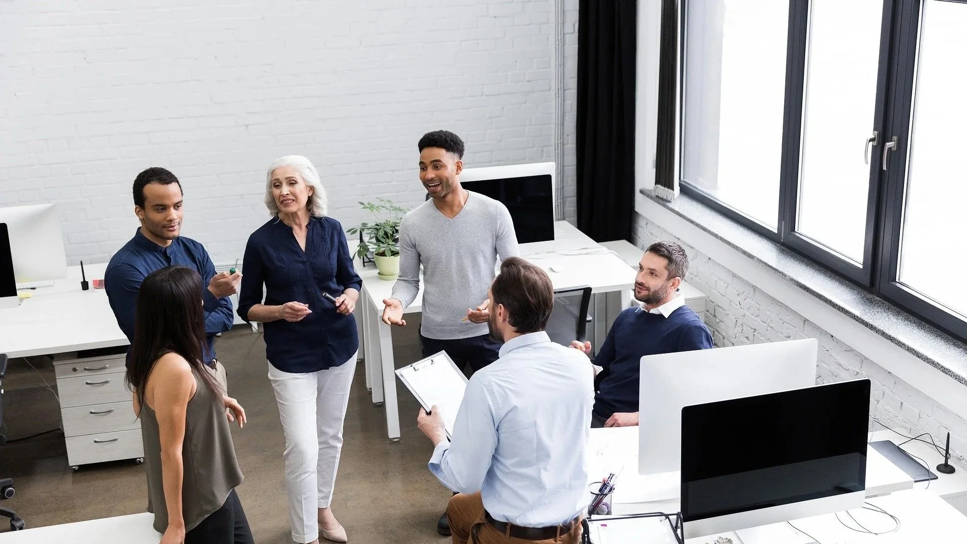 Group of people in a modern office, engaged in a discussion or meeting. Five seated and standing individuals, with two women and three men, appear attentive and engaged.