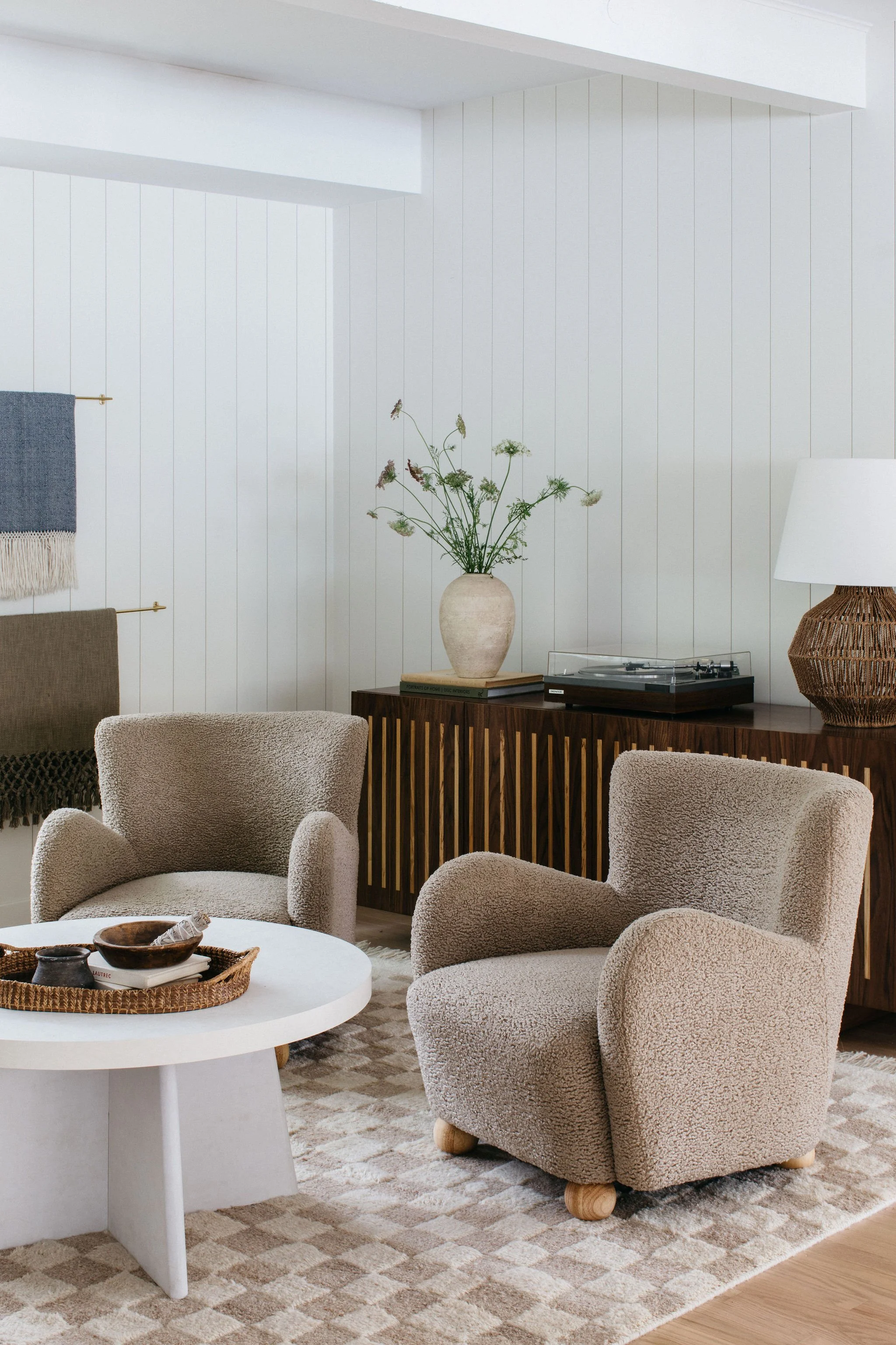 Cozy living room with beige armchairs, a white round coffee table with decorative bowls, a beige patterned rug, a wooden sideboard with a flower vase and books, and a white wall with a hanging textile, a lamp with a woven base, and a record player on the sideboard.