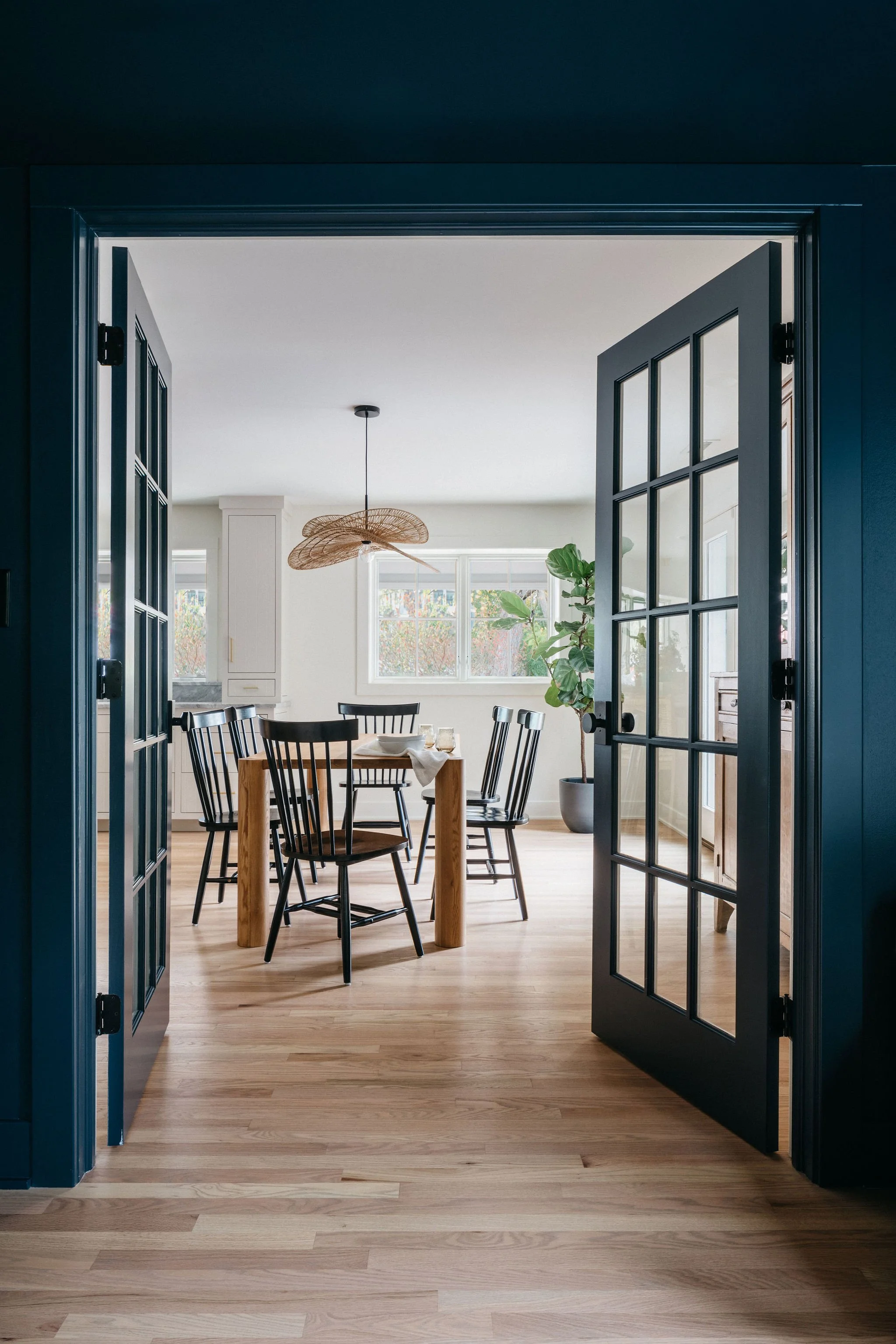 View of a cozy dining room through blue double glass paneled doors, featuring a wooden table surrounded by black chairs, a large window with natural light, a hanging wicker pendant light, and a potted plant.