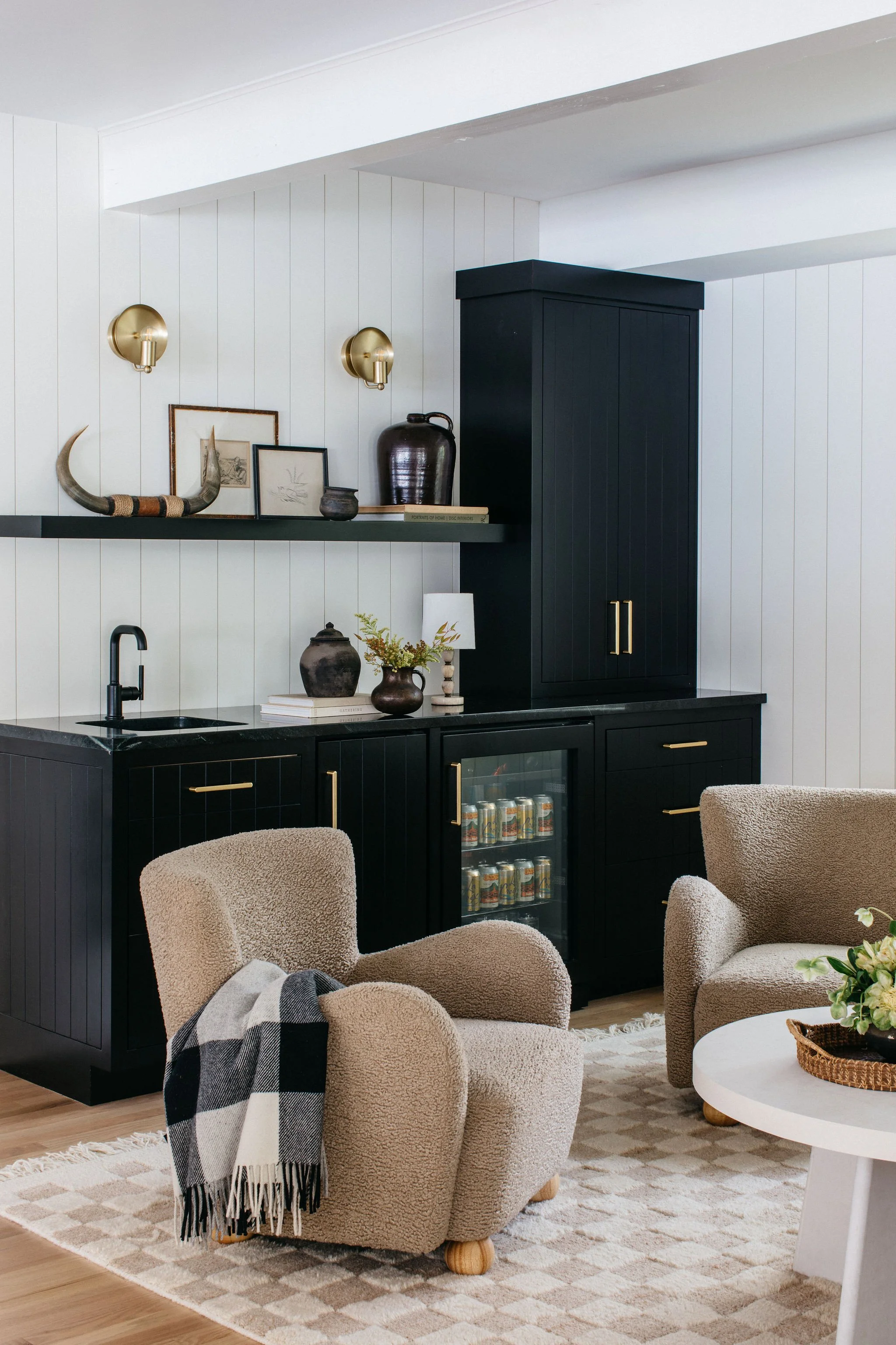 Cozy living room area featuring a black cabinet with gold handles, beige armchairs, a patterned rug, and decorative items on shelves, including framed artwork, a large dark pottery vase, and a white table with a wicker tray.
