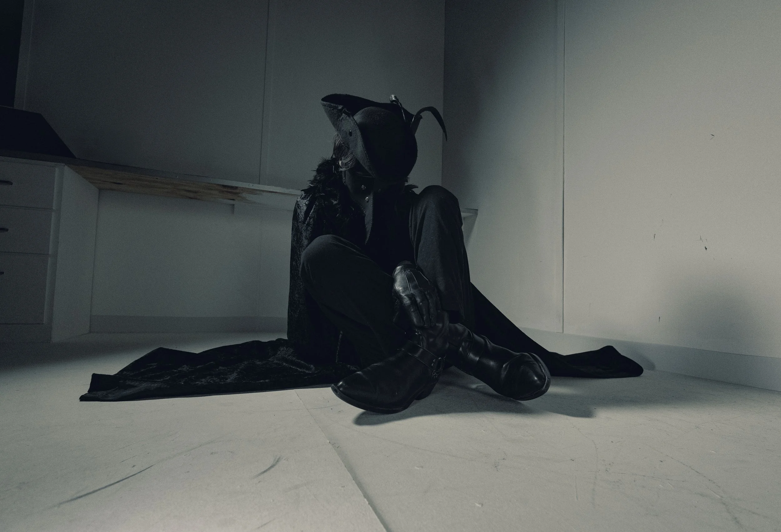 Person dressed in black sits on the floor in a minimalist, gray room, with their head resting to the side and wearing a large hat. The person appears to be leaning against the wall, with their legs crossed and hands resting on their knee.