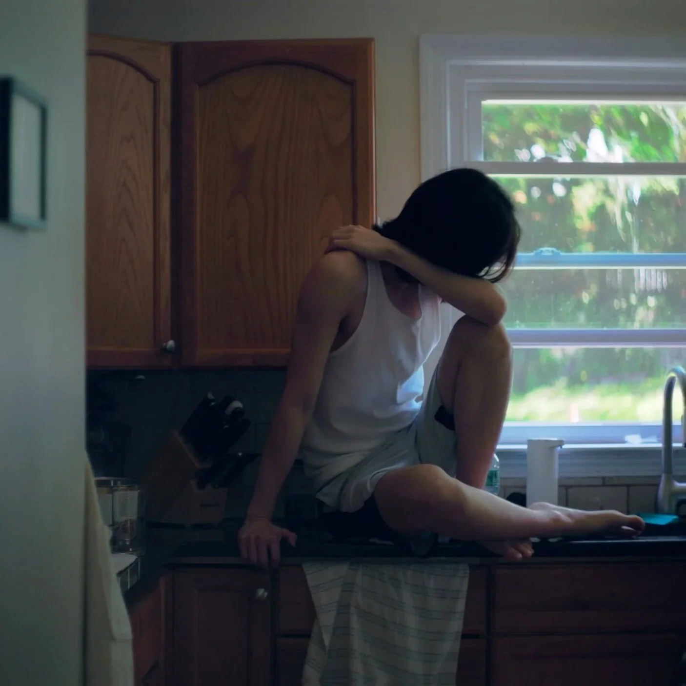 Person sitting on kitchen counter with one knee up near window, wearing a white tank top and shorts, leaning forward with head resting on arm.