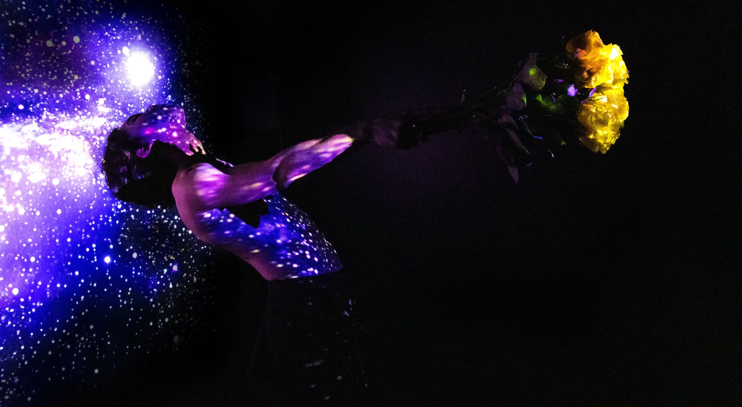 A person is lying on a dark floor, illuminated by blacklight, along with a bouquet of yellow flowers. The person and the flowers glow under the UV light, with the person's body showing purple, blue, and white reflections.