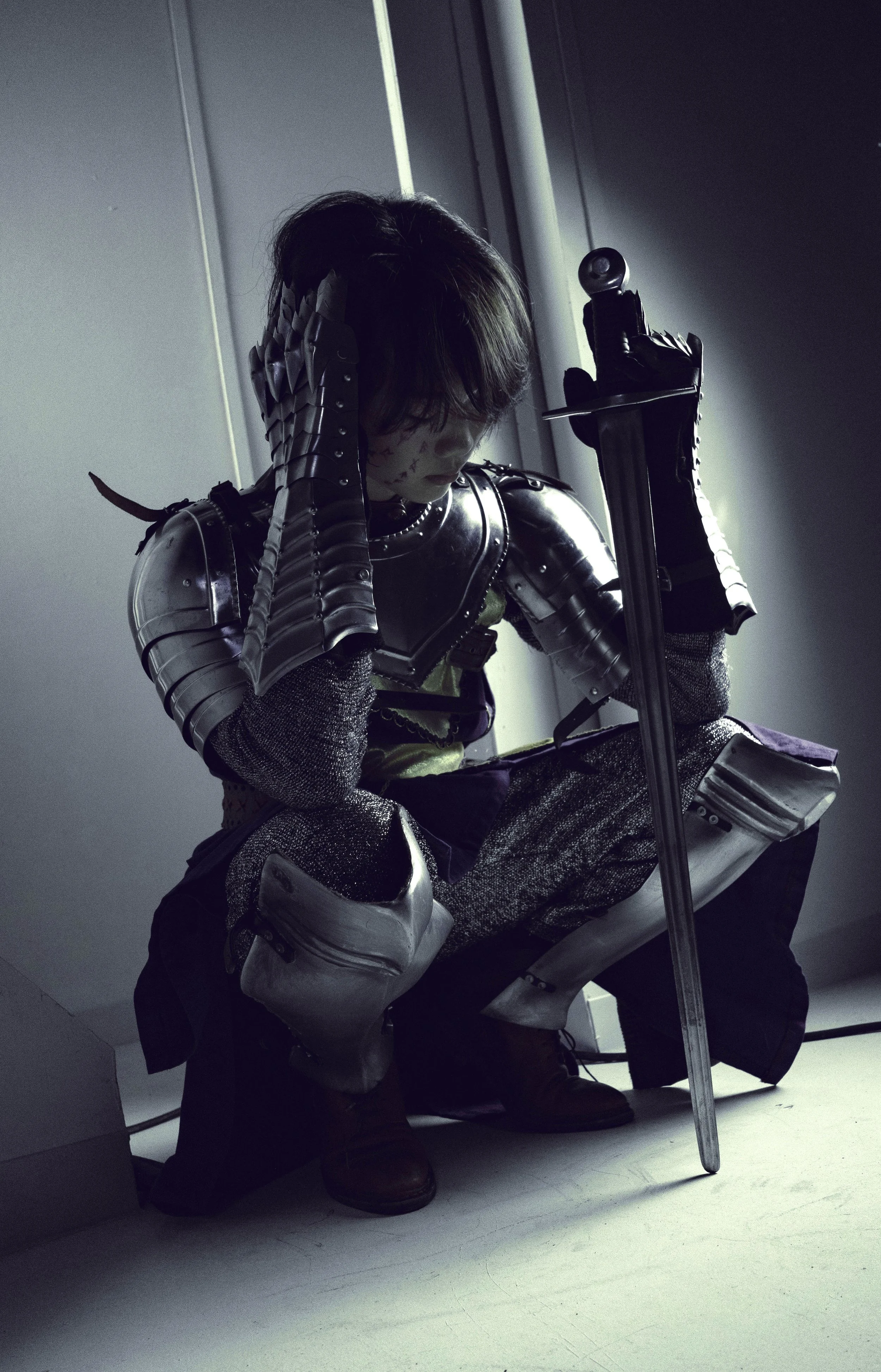 Person dressed as a medieval knight in armor sitting on a chair and holding a sword, with dark lighting and a modern indoor setting.