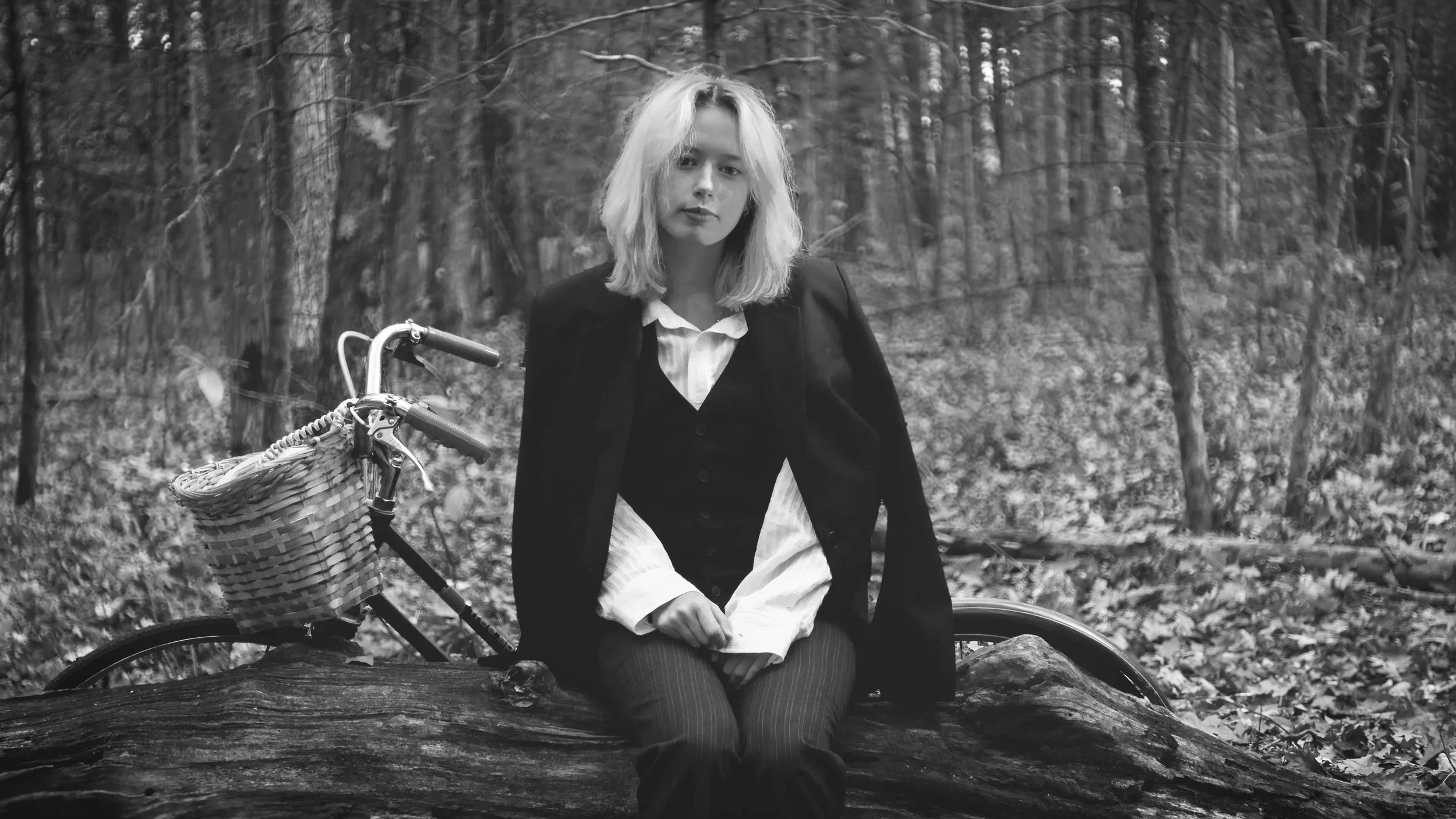 A woman with light-colored hair in a forest setting, sitting on a log, with a bicycle beside her that has a wicker basket attached to the handlebars.