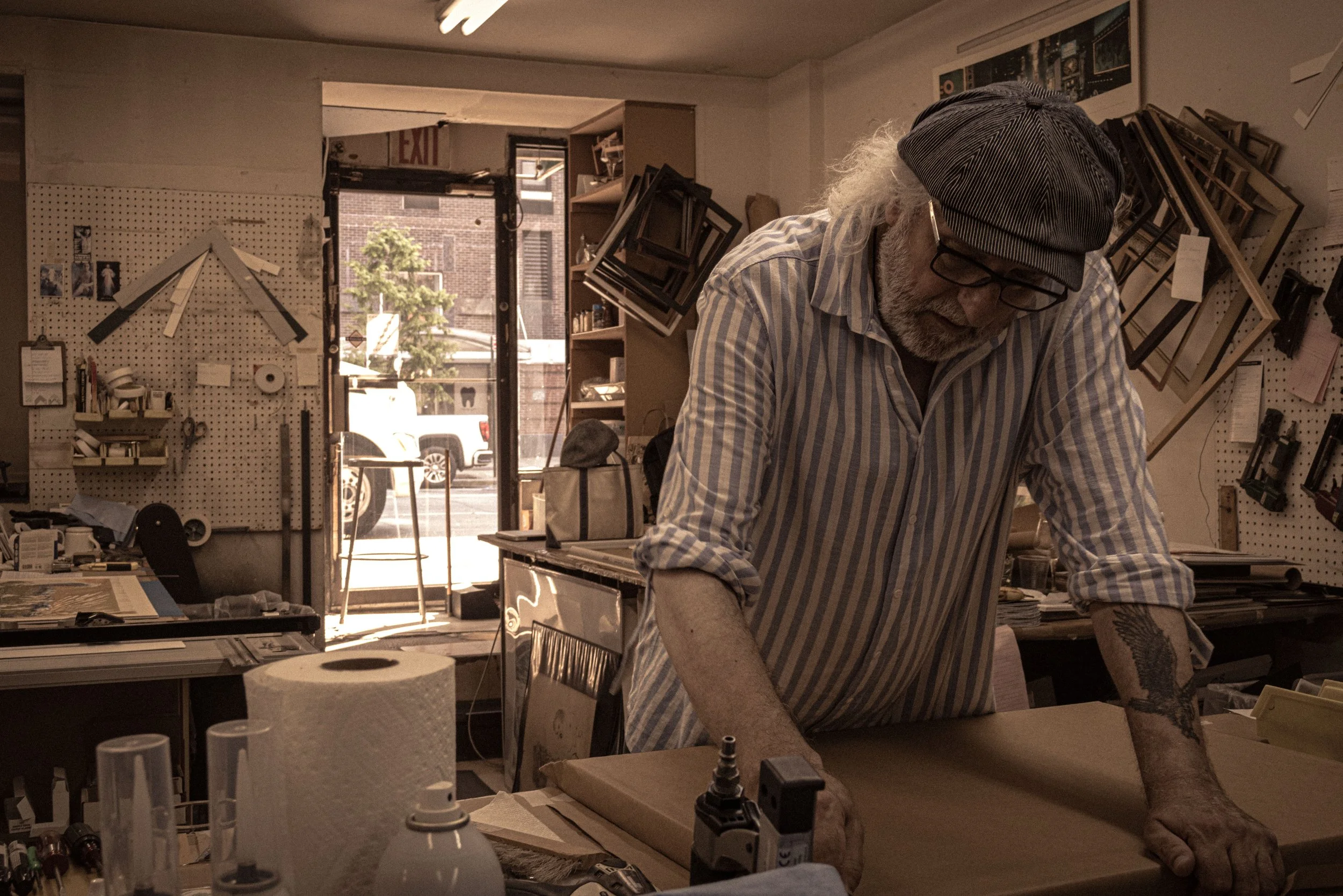 An elderly man with white hair and a beard wearing a striped shirt, a cap, and glasses working in a cluttered workshop. The background shows pegboards with tools, framed pictures, and a computer monitor, with a glass door revealing a street scene out