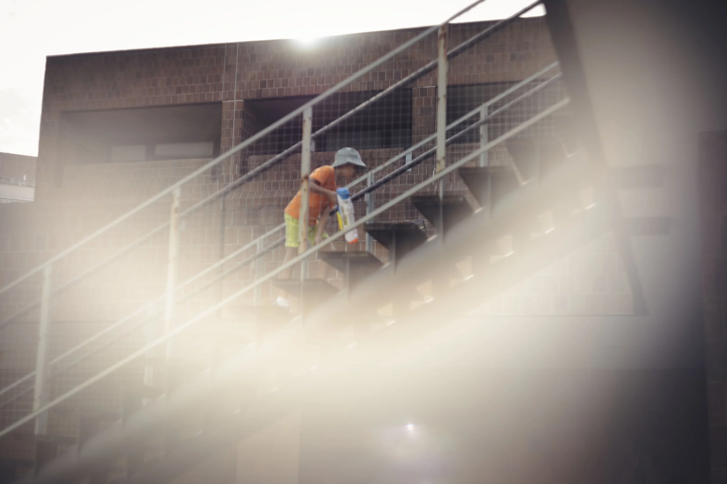 A young child in a hat and colorful clothing is climbing up an outdoor metal staircase attached to a brick building, with the sun shining in the background.