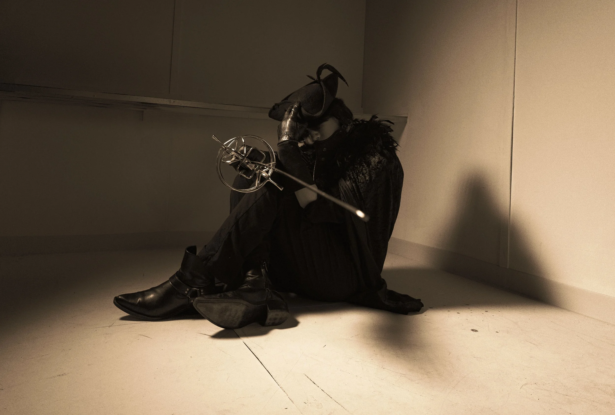 A person dressed in black, sitting on the floor, holding a sword, with their head bowed and hand on forehead, in a dimly lit room.