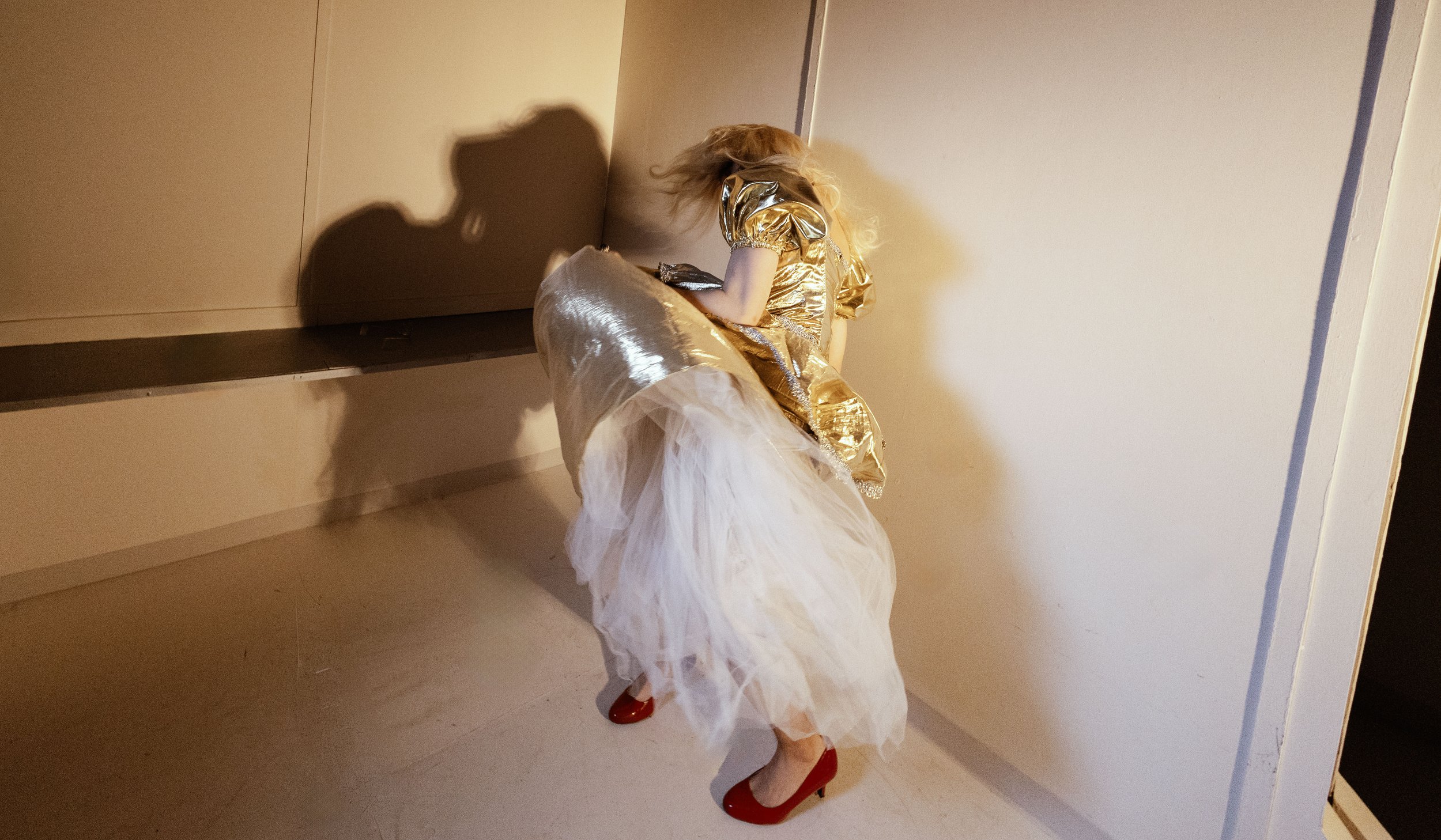 A young girl dressed in a golden top, white tulle skirt, and red shoes, standing against a wall, with her face turned away from the camera.