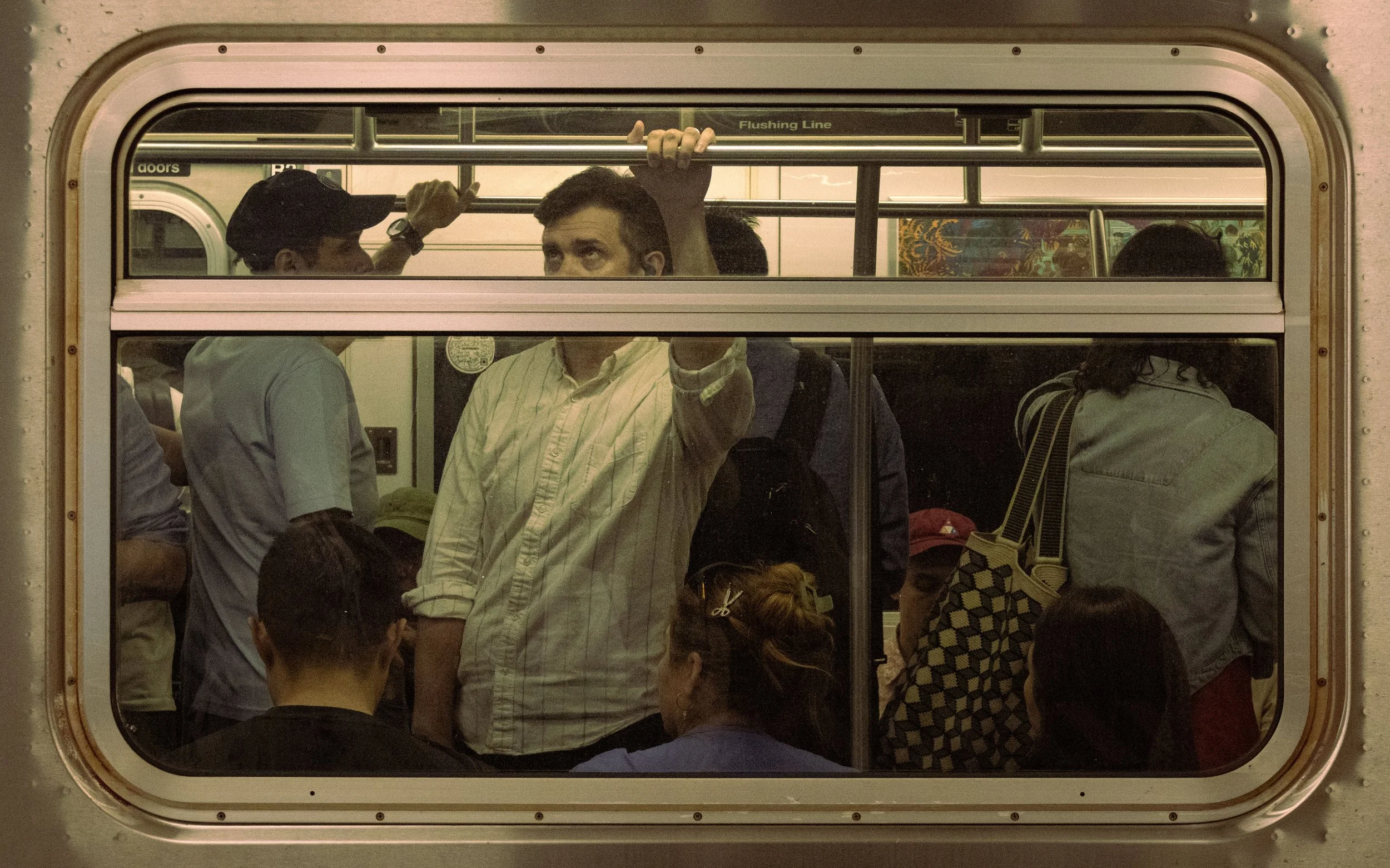 Several people standing and sitting on a bus, viewed through a rectangular bus window. Some are facing the window, including a man with a backpack and a woman with curly hair. Others are looking away or obscured.
