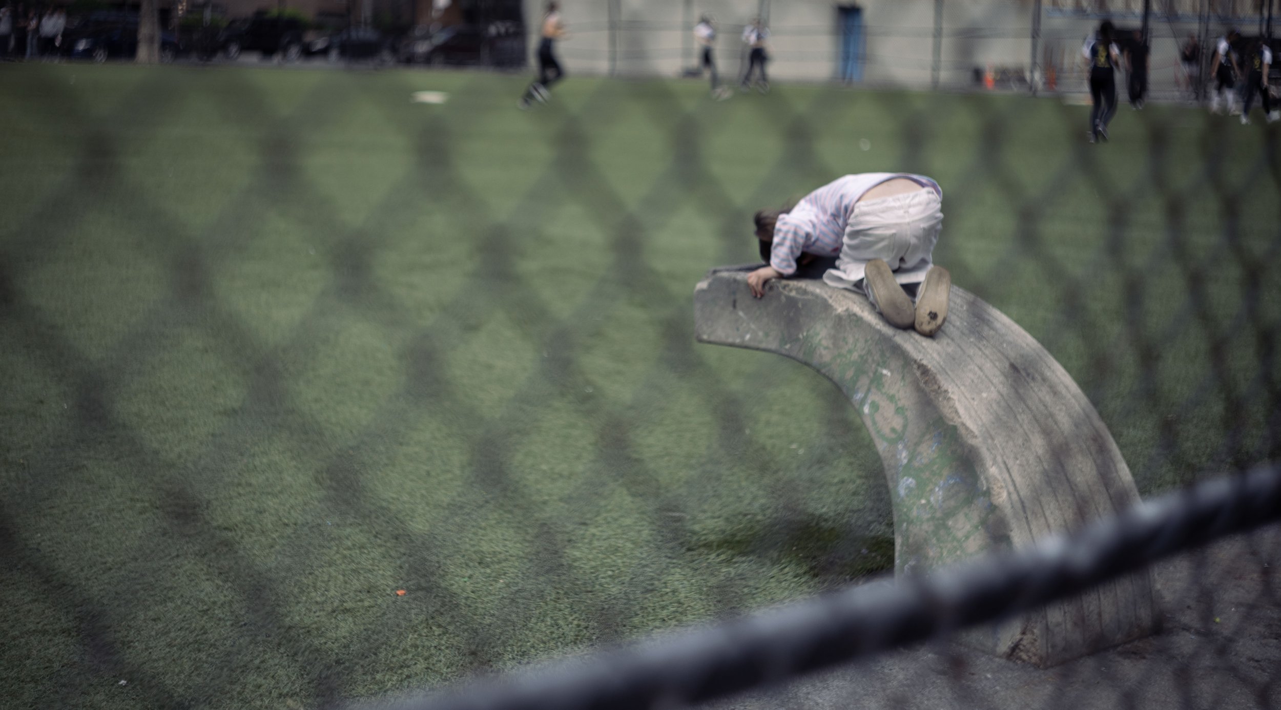 Person kneeling on a skate ramp, reaching down with one hand, at a skate park with other skaters in the background.