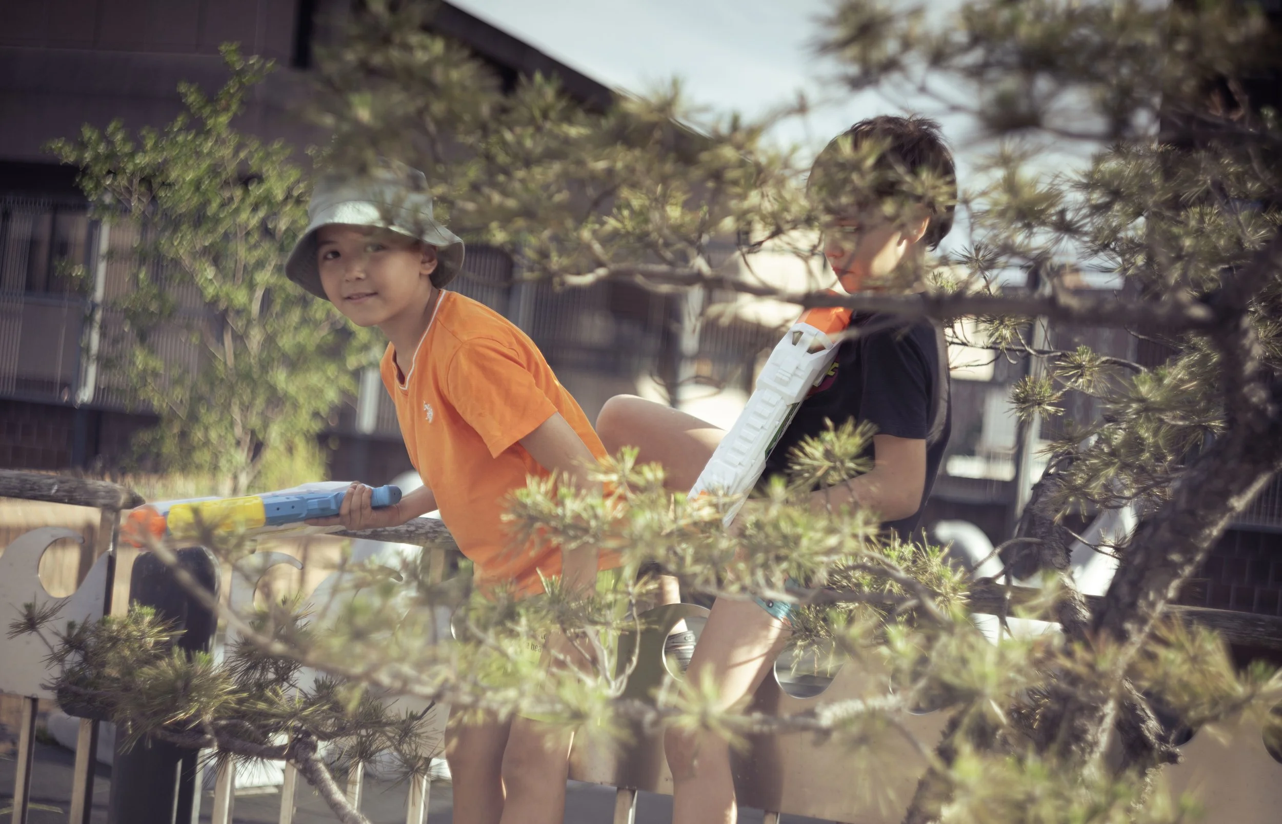 Two children playing outside near a tree, one boy with a bucket hat and orange shirt, and another boy with glasses and black shirt, holding a water gun.
