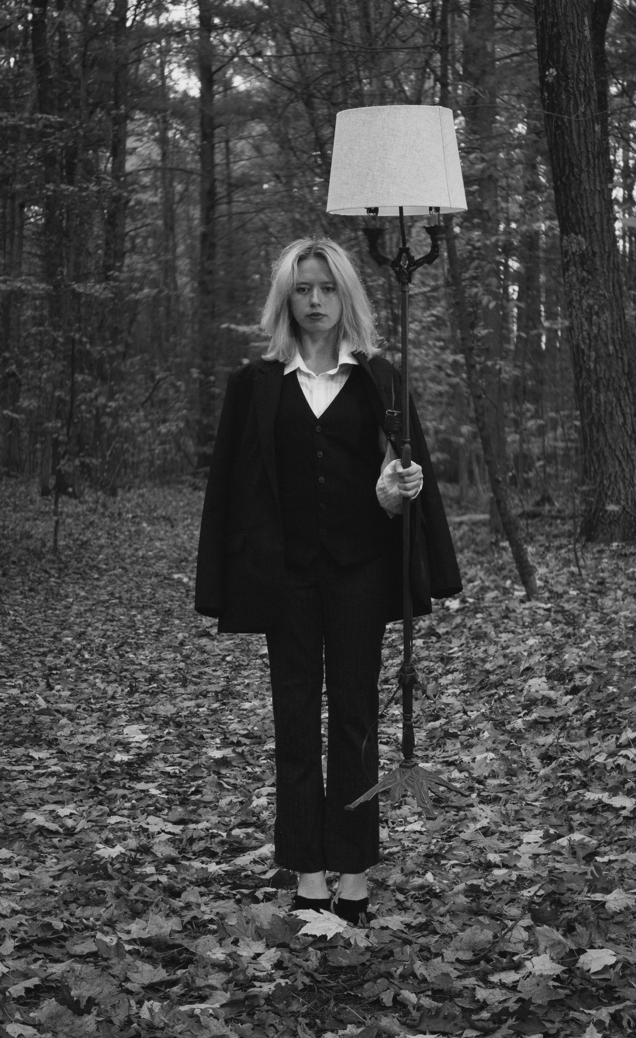 A woman with shoulder-length blonde hair stands in a forest, holding an umbrella with a lamp shade on top. She is dressed in a dark suit with a white shirt and appears serious. The scene is in black and white, with leaves covering the ground and tree