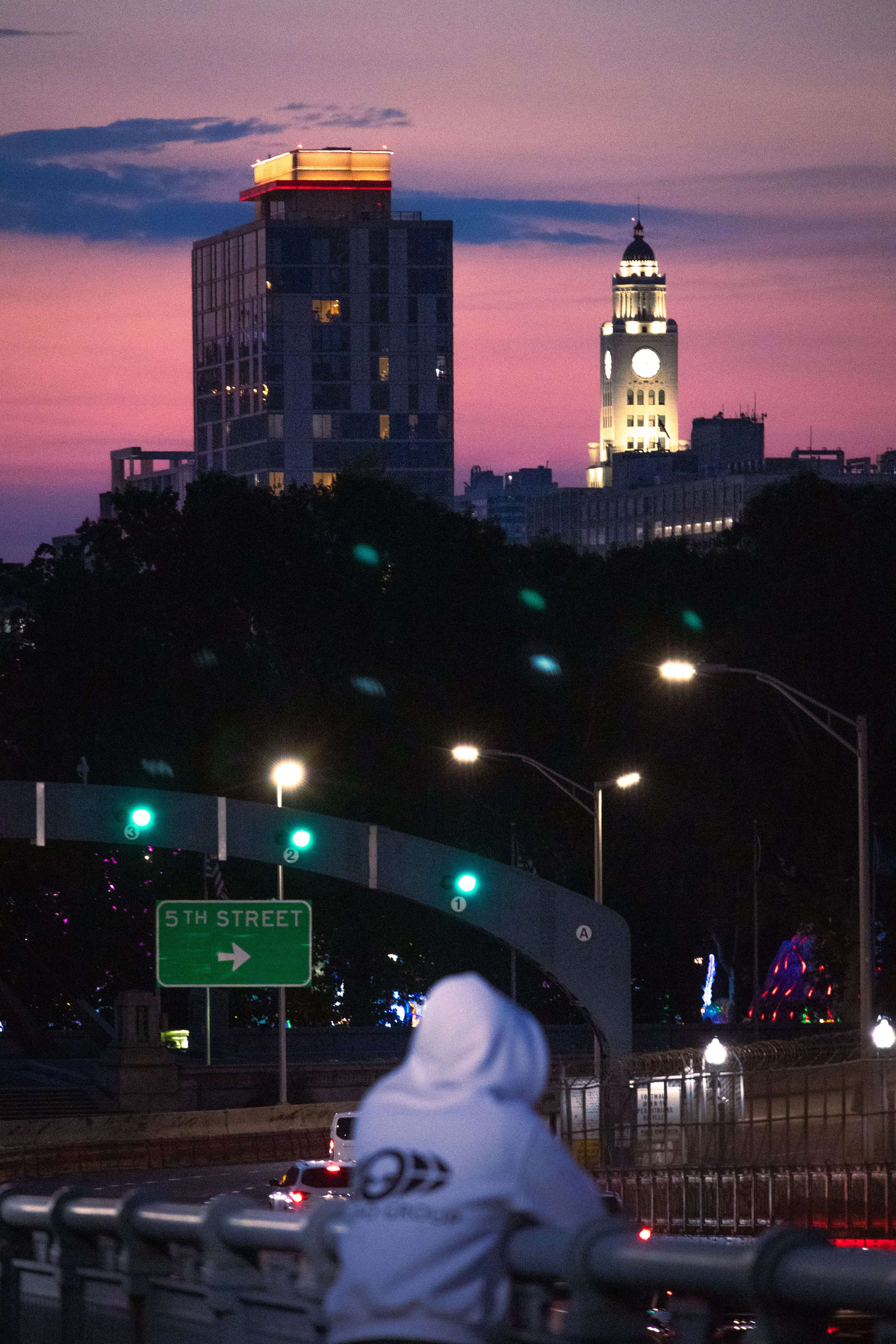 A city skyline at dusk with a tall clock tower and a modern high-rise building, pink and purple sunset sky, street lights, cars, and a green street sign for 5th Street.