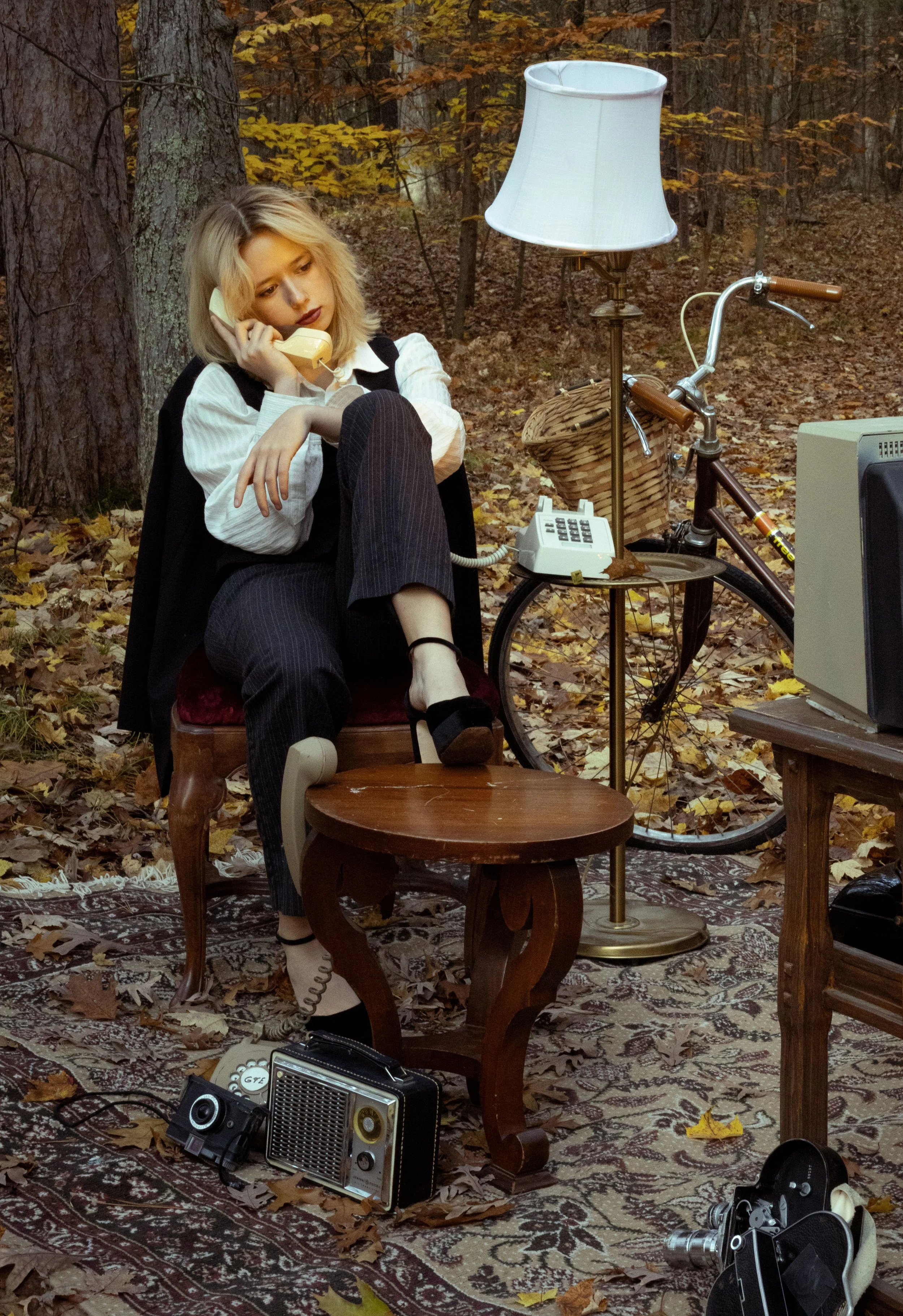 A woman with blonde hair, dressed in a white blouse and black pants, seated in a forest surrounded by fallen autumn leaves, talking on a yellow rotary phone. She has a serious expression. Nearby, a vintage bicycle with a wicker basket is visible, and