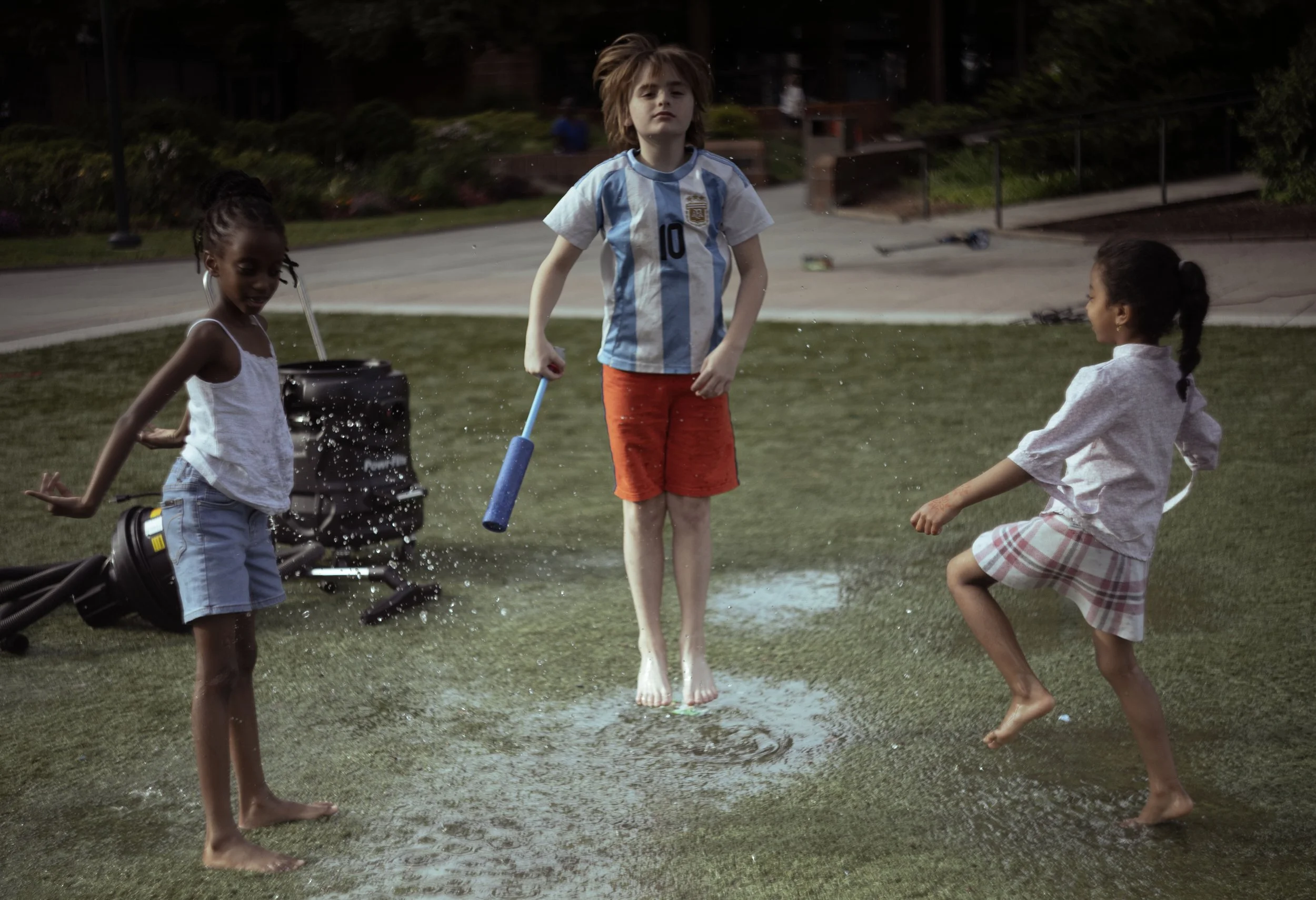 Children playing on a water mat in a park during daytime, with trees and a pathway in the background.