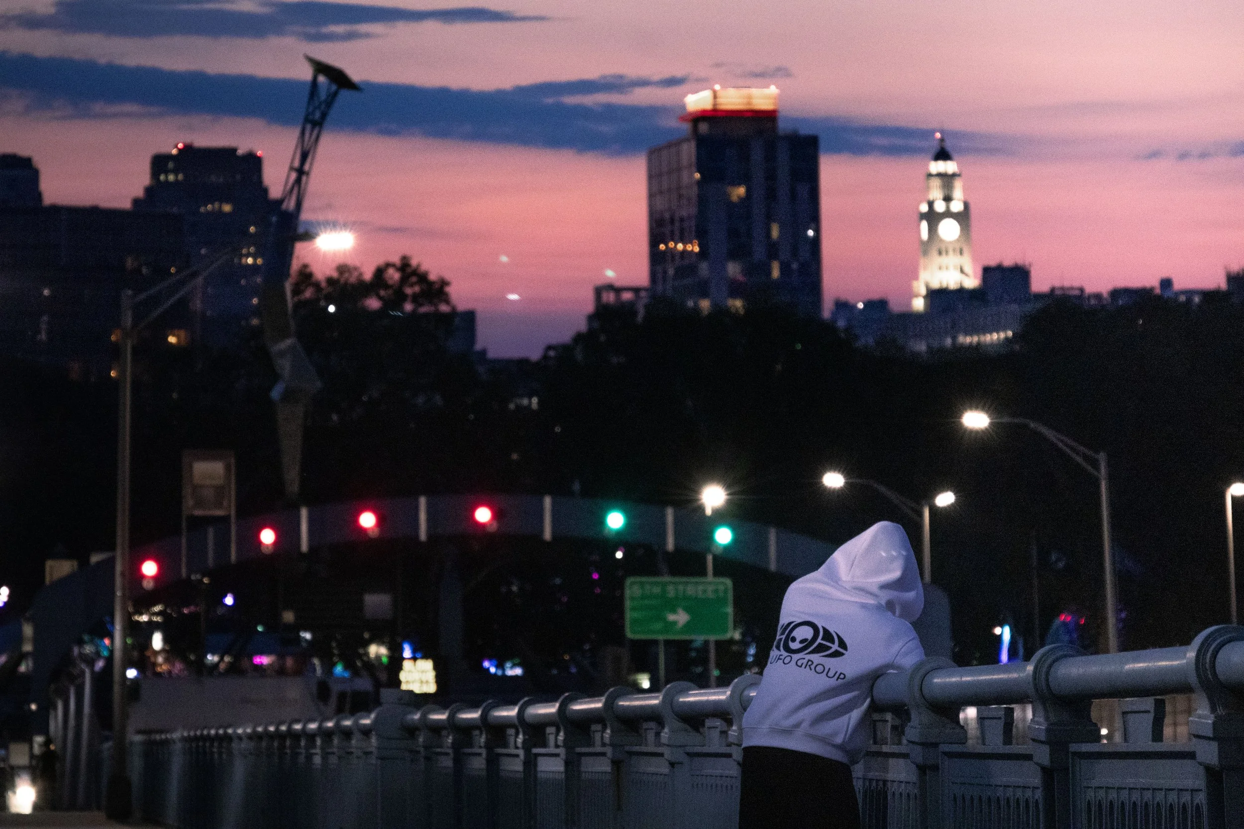 City skyline at dusk with tall buildings, including a clock tower, under a colorful sky. In the foreground, a person wearing a white jacket is leaning on a railing near a road with red and green traffic lights.