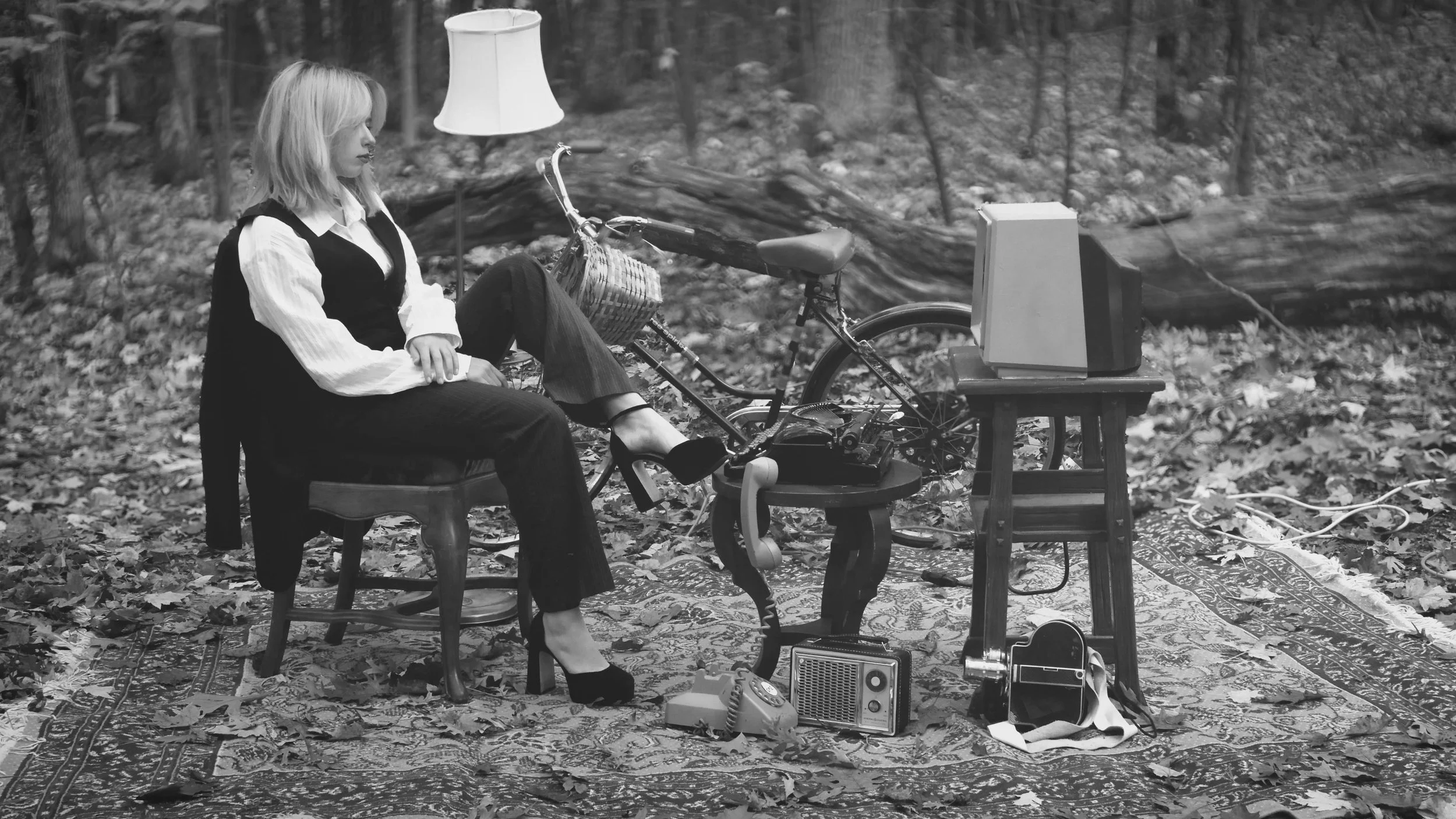A woman sitting outdoors on a wooden chair next to a bicycle, with a lamp, a table, and vintage electronic devices including a radio and a television set in front of her. The ground is covered with fallen leaves, and there is a patterned rug beneath 