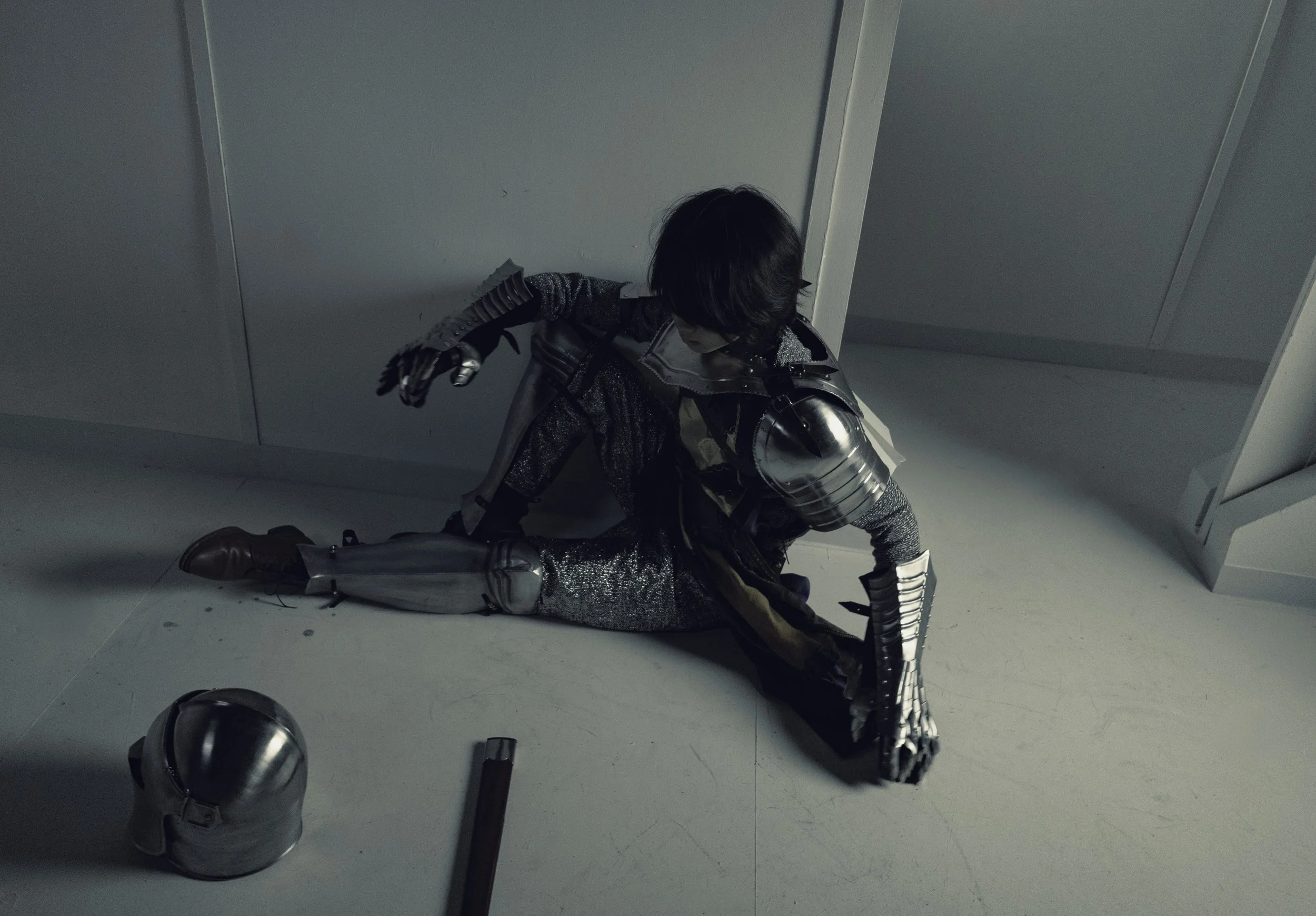 A person dressed in an armored costume, black hair, kneeling on the ground, with a metallic helmet and gloves, in a gray room with debris.