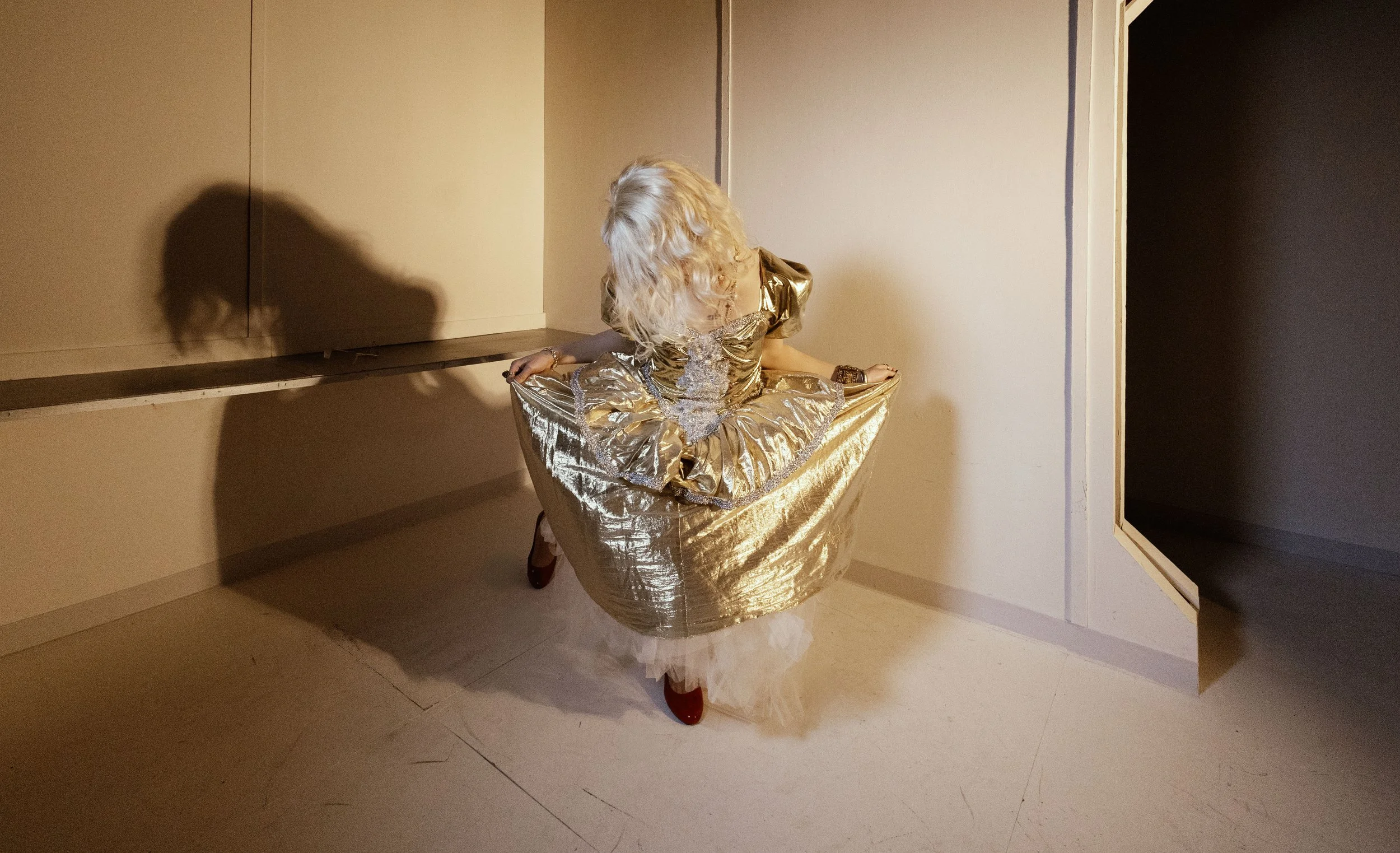A person with blonde curly hair is sitting inside a large, gold-colored, metallic-looking bowl with a wide rim. The person is wearing a matching gold dress with puffed sleeves and is looking downward or away, with their head bent and face not visible