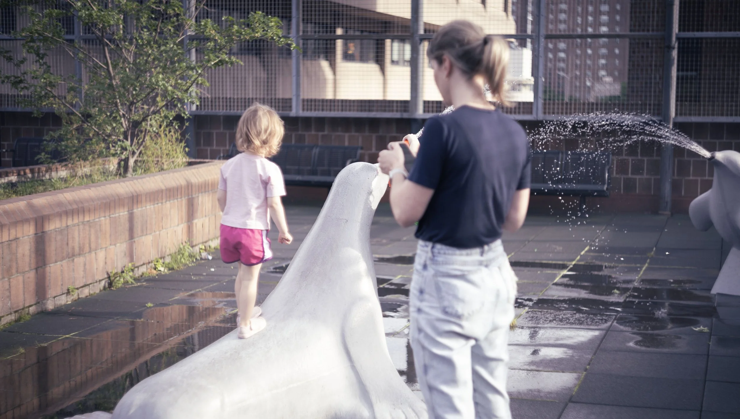 Two children, a girl and a boy, playing at a rooftop playground with animal-shaped water fountains, with water spraying from the fountains.