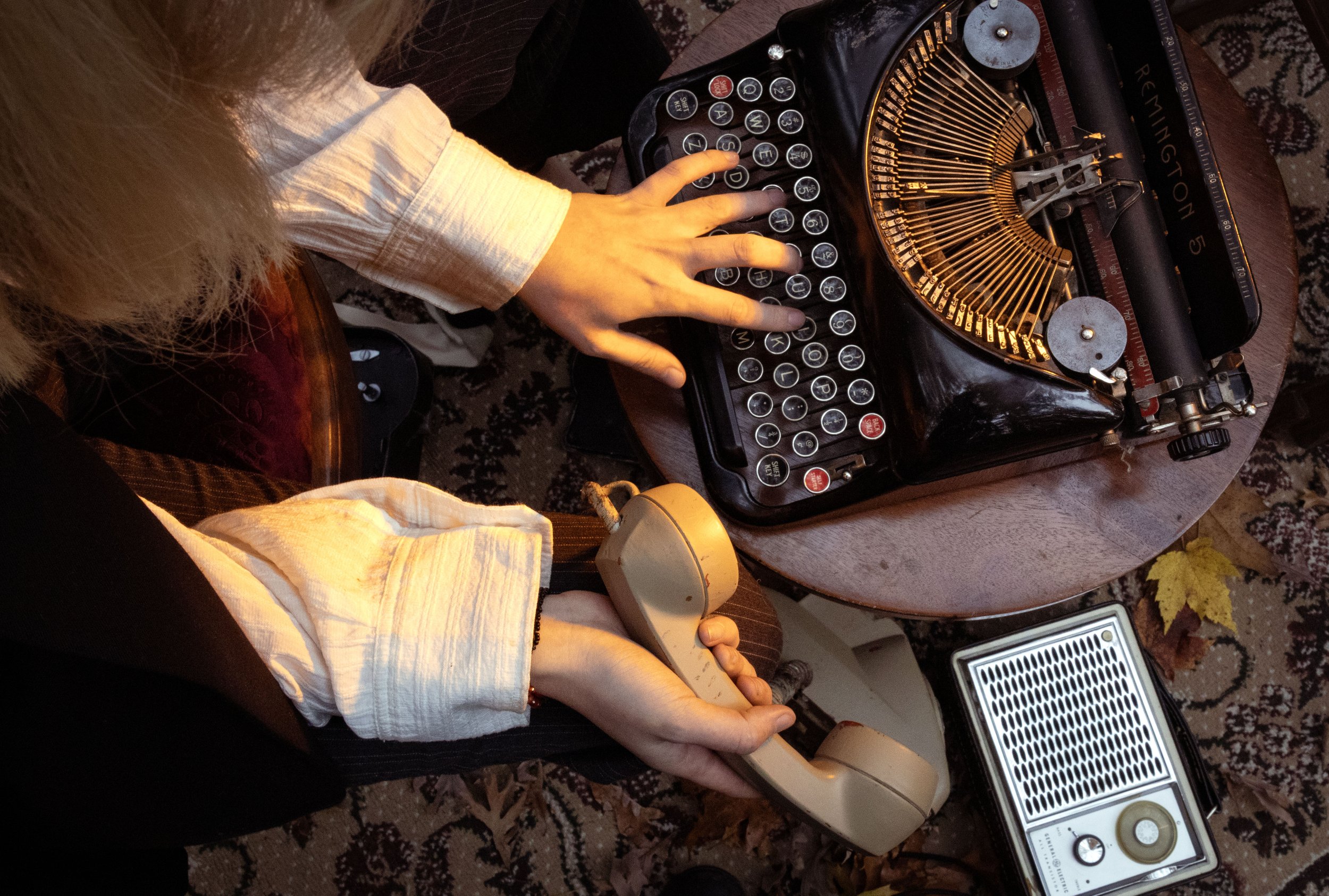 A person's hand resting on a vintage typewriter, with a computer keyboard and speakers nearby, on a wooden table with a patterned rug underneath. The person is wearing a light-colored sleeve, and part of their hair and torso are visible.