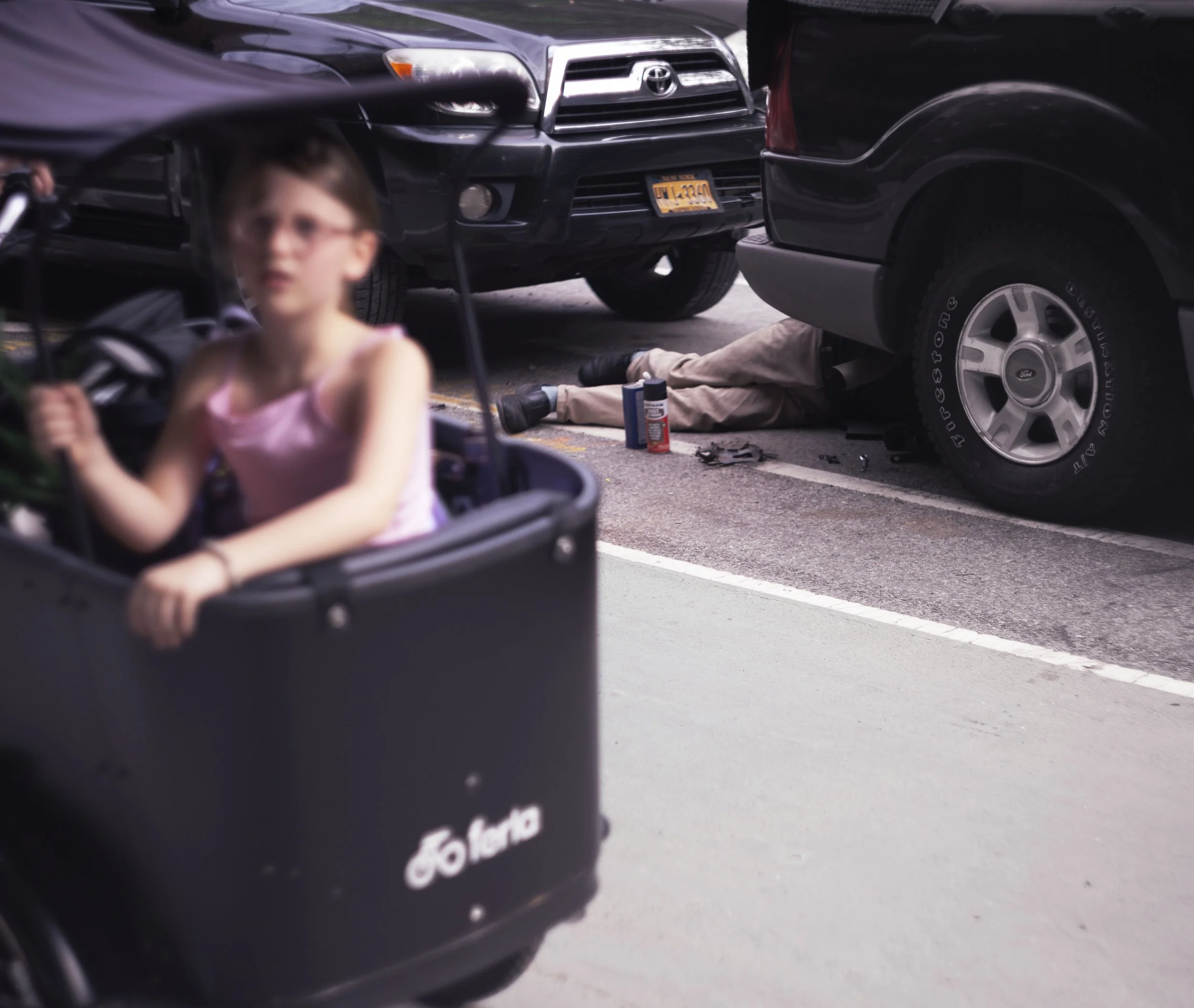 Blurred image of a young girl in a stroller in the foreground with cars parked on the street behind her, including a Toyota and a Ford. Someone is lying underneath the front of a black SUV, with only their legs visible. Tools and car maintenance supp