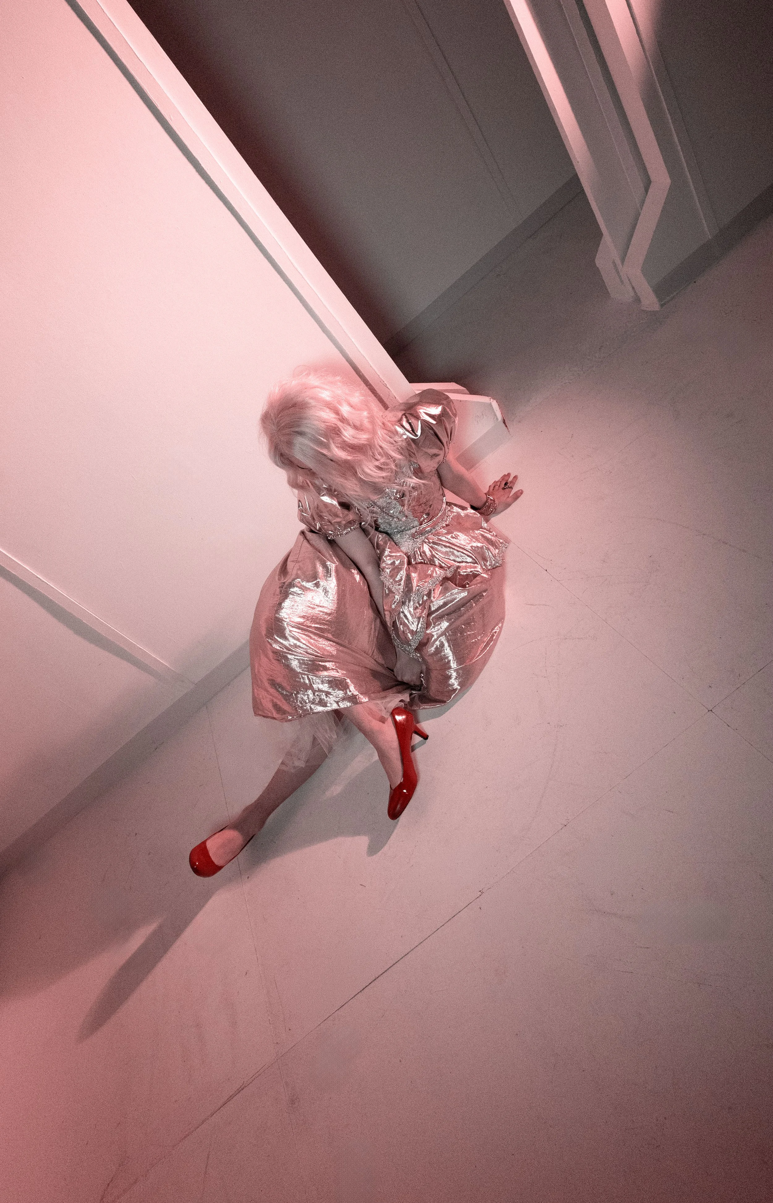 A person dressed in a shiny, metallic silver and pink dress with matching red high heels, sitting on a pink bench against a plain pink wall with a matching pink floor.