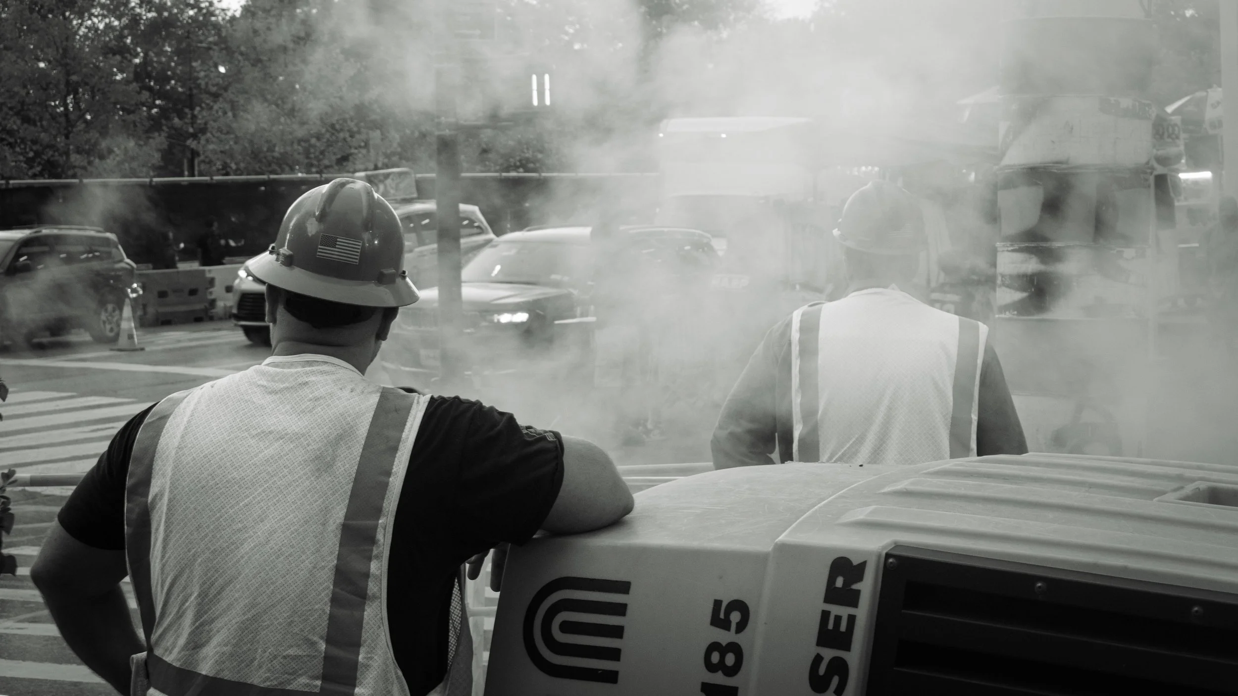 Two workers wearing safety helmets and reflective vests are working near a street, with smoke or dust in the air, and cars passing by in the background.