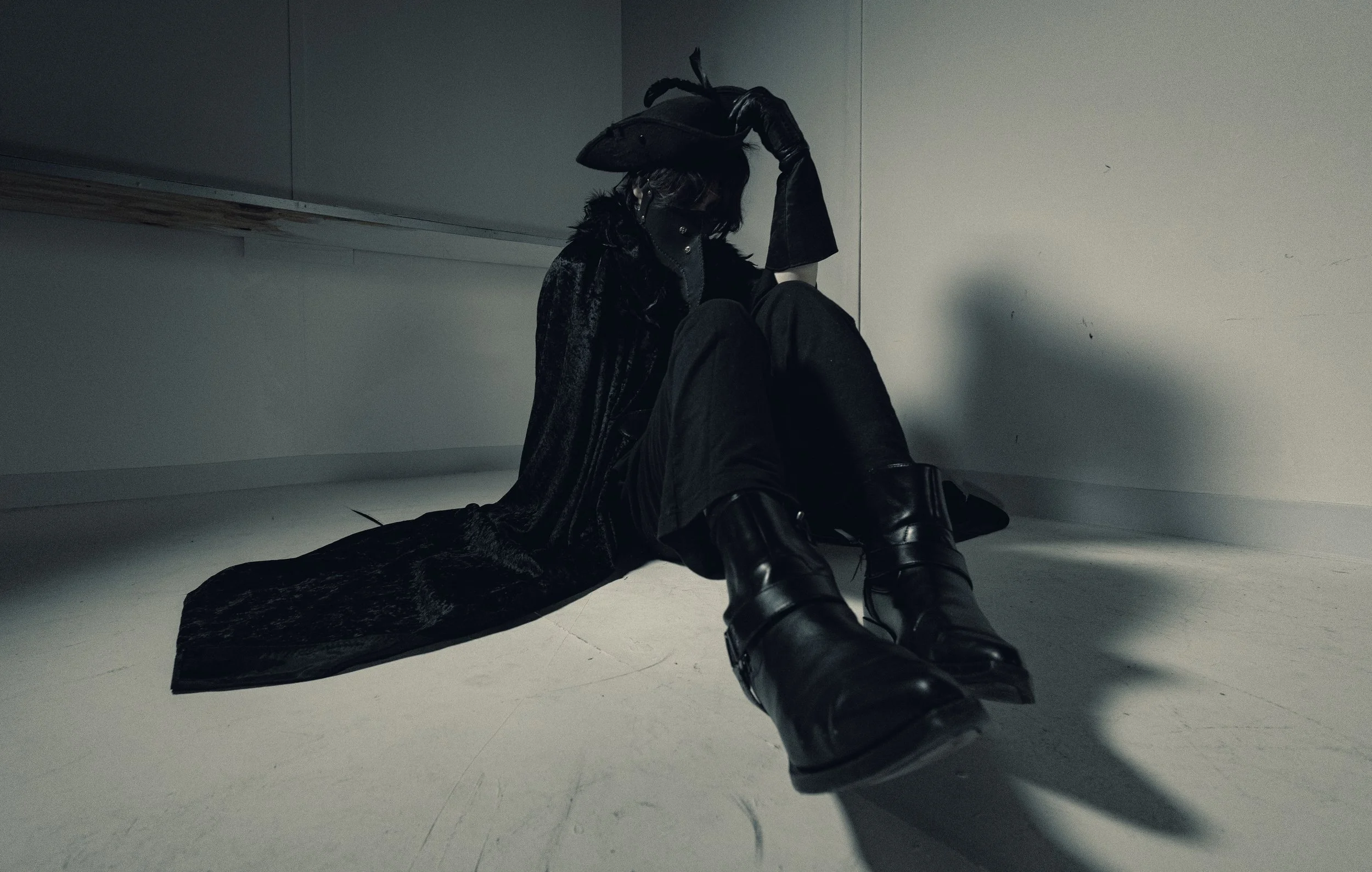 Person sitting on the floor in a corner, wearing black leather gloves and boots, with a patterned black coat, with their face partially obscured, in a dimly lit room with plain walls and a reflective floor.